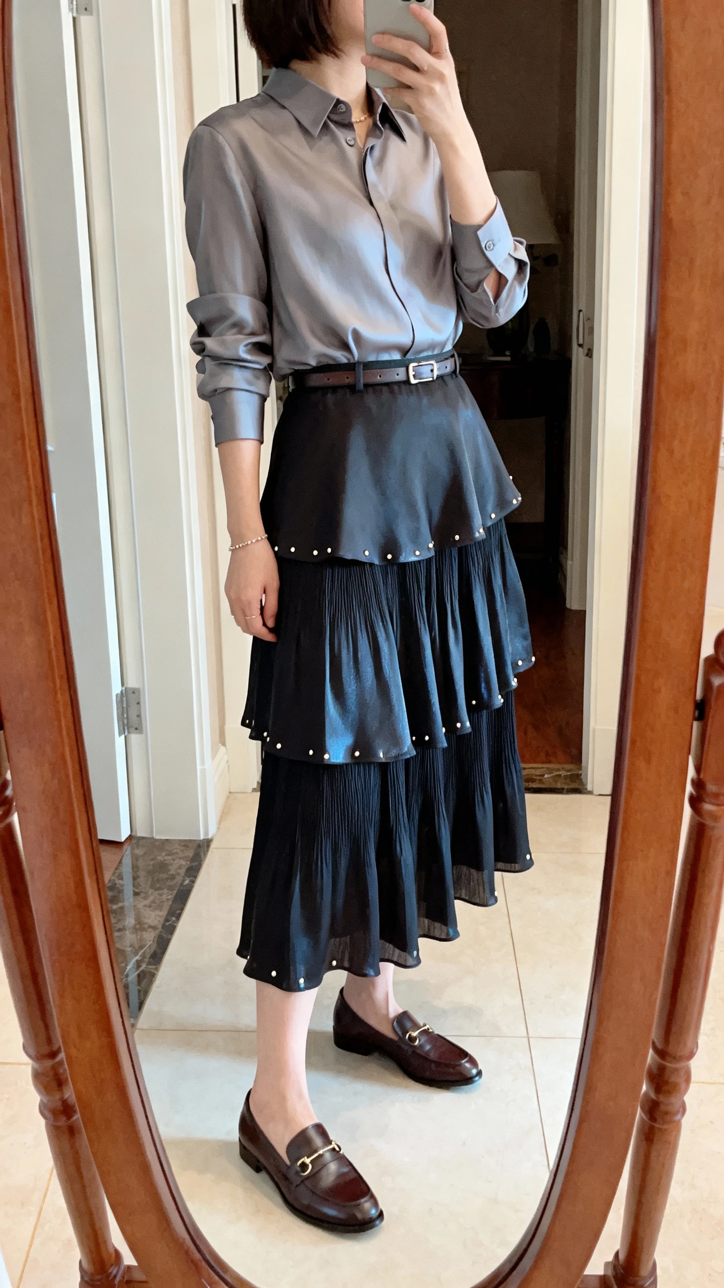 iPhone mirror selfie of a woman wearing a polished shirt-blouse tucked into a tiered skirt with a slight sheen, slim belt, dainty studs, and classic loafers, face not visible, side angle, full-length mirror in entryway, natural indoor light, iPhone photo quality