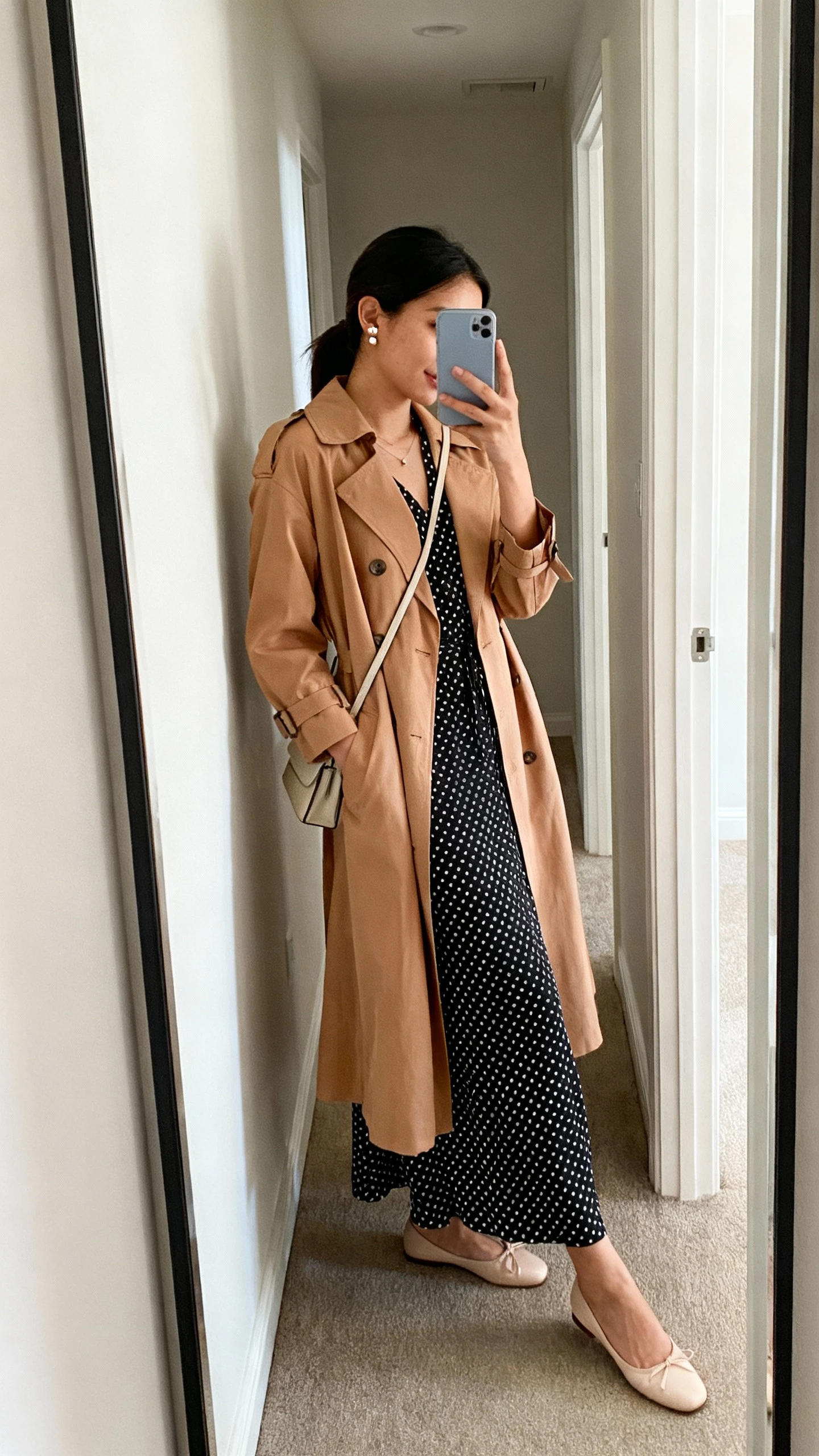 iPhone mirror selfie of a woman wearing a polka dot maxi dress under a lightweight tan trench coat, ballet flats, delicate stud earrings, simple crossbody bag, face not visible, side angle in a hallway full-length mirror, soft natural daylight, iPhone photo quality