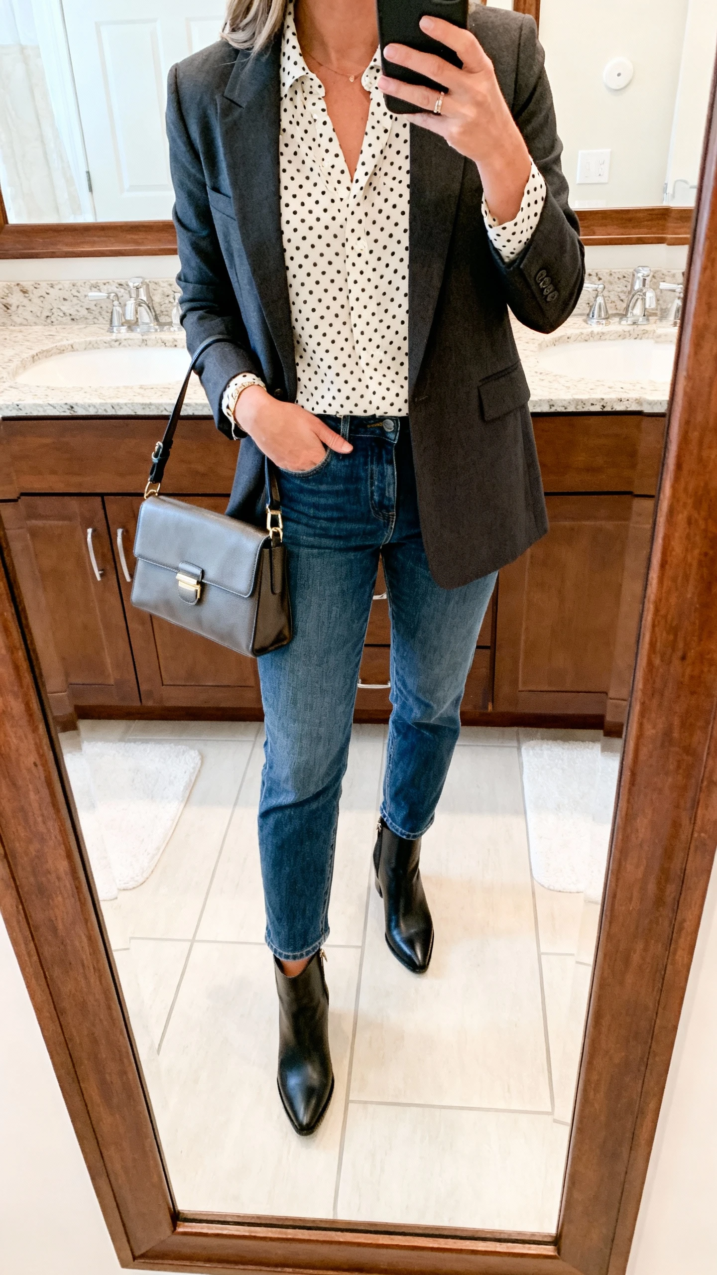 iPhone mirror selfie of a woman wearing a polka-dot micro-print blouse layered under a tailored blazer with straight-leg jeans, ankle boots, and a structured satchel, face not visible, cropped at neck, bathroom mirror, bright indoor lighting, iPhone photo quality.