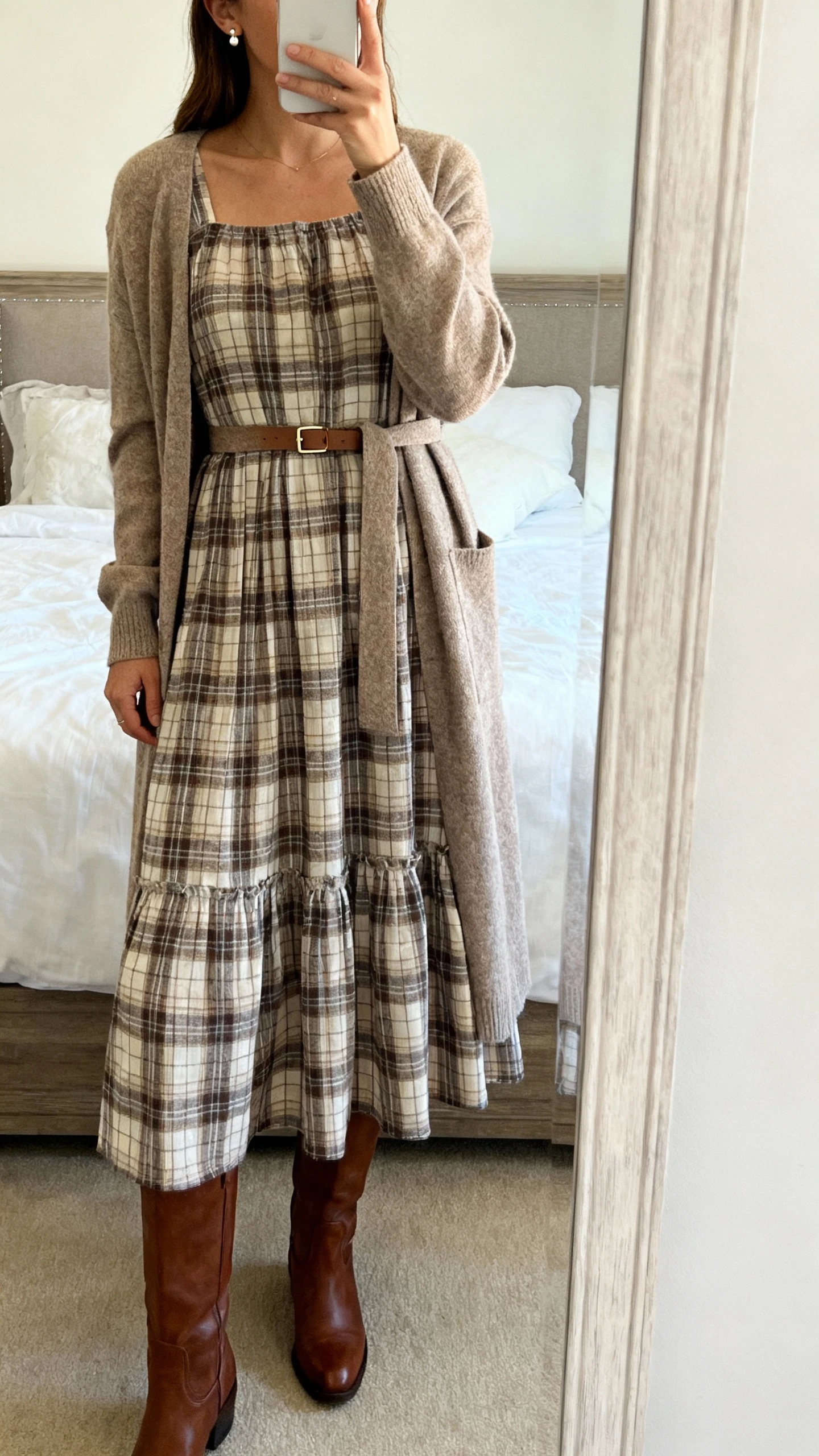 iPhone mirror selfie of a woman wearing a prairie-inspired plaid midi dress with a belted longline cardigan, tall leather boots, a slim waist belt, and delicate stud earrings, face not visible, cropped at shoulders, bedroom mirror, soft daylight, iPhone photo quality.