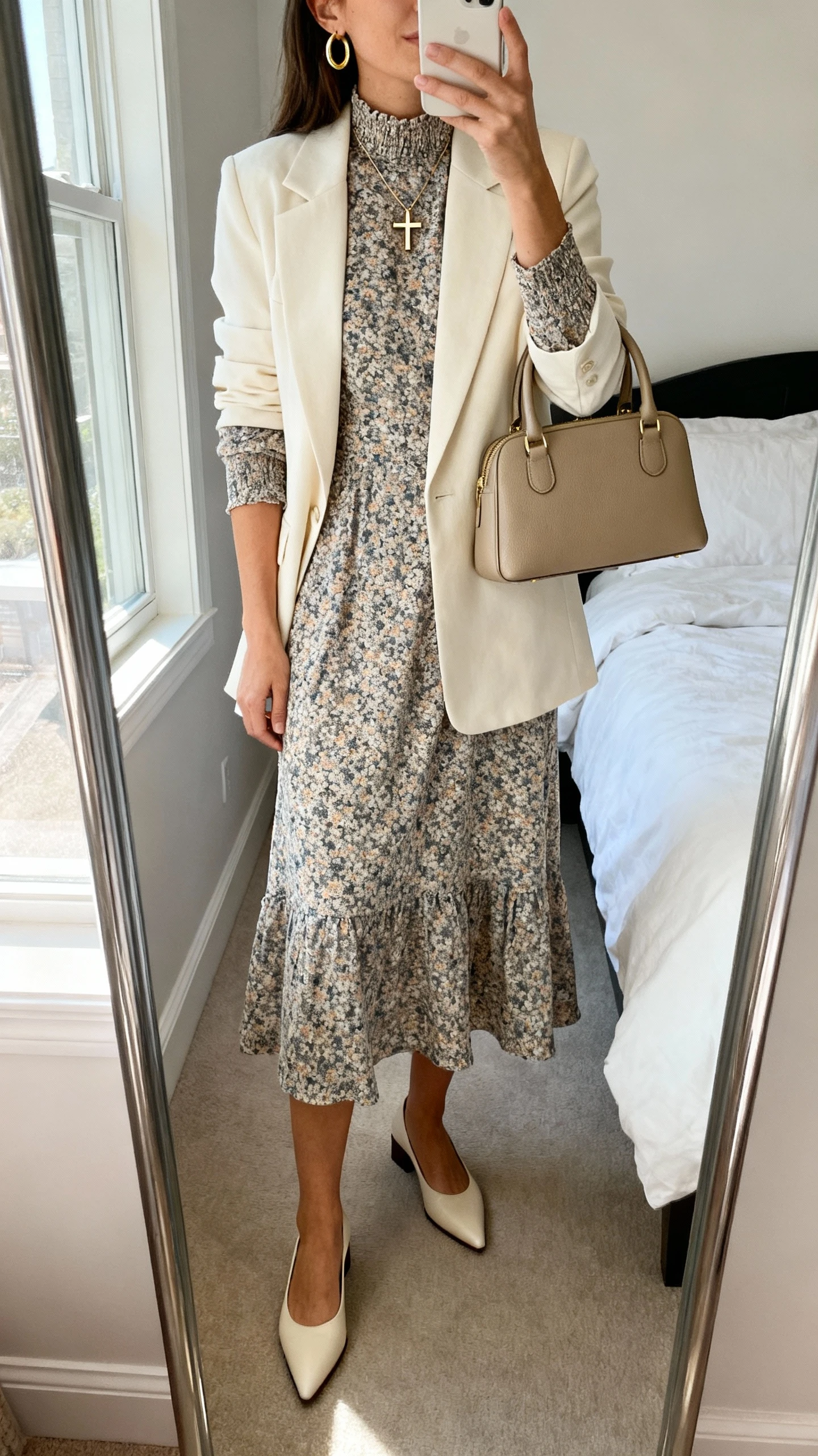 iPhone mirror selfie of a woman wearing a printed midi dress with a high neckline and elbow sleeves, a lightly structured cream blazer, pointed-toe low block heels, minimal gold hoop earrings, a delicate cross pendant, and a top-handle bag; face not visible, cropped at neck, bedroom mirror, natural window light, iPhone photo quality.