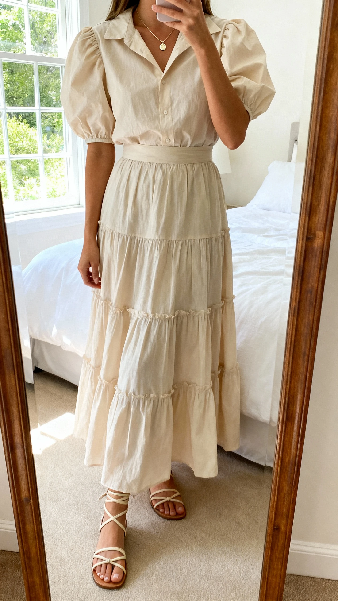 iPhone mirror selfie of a woman wearing a puff-sleeve poplin blouse tucked into a tiered maxi skirt for modern romance, accessorized with a dainty pendant necklace and strappy sandals, face not visible, cropped at shoulders, bedroom mirror, bright window light, iPhone photo quality.