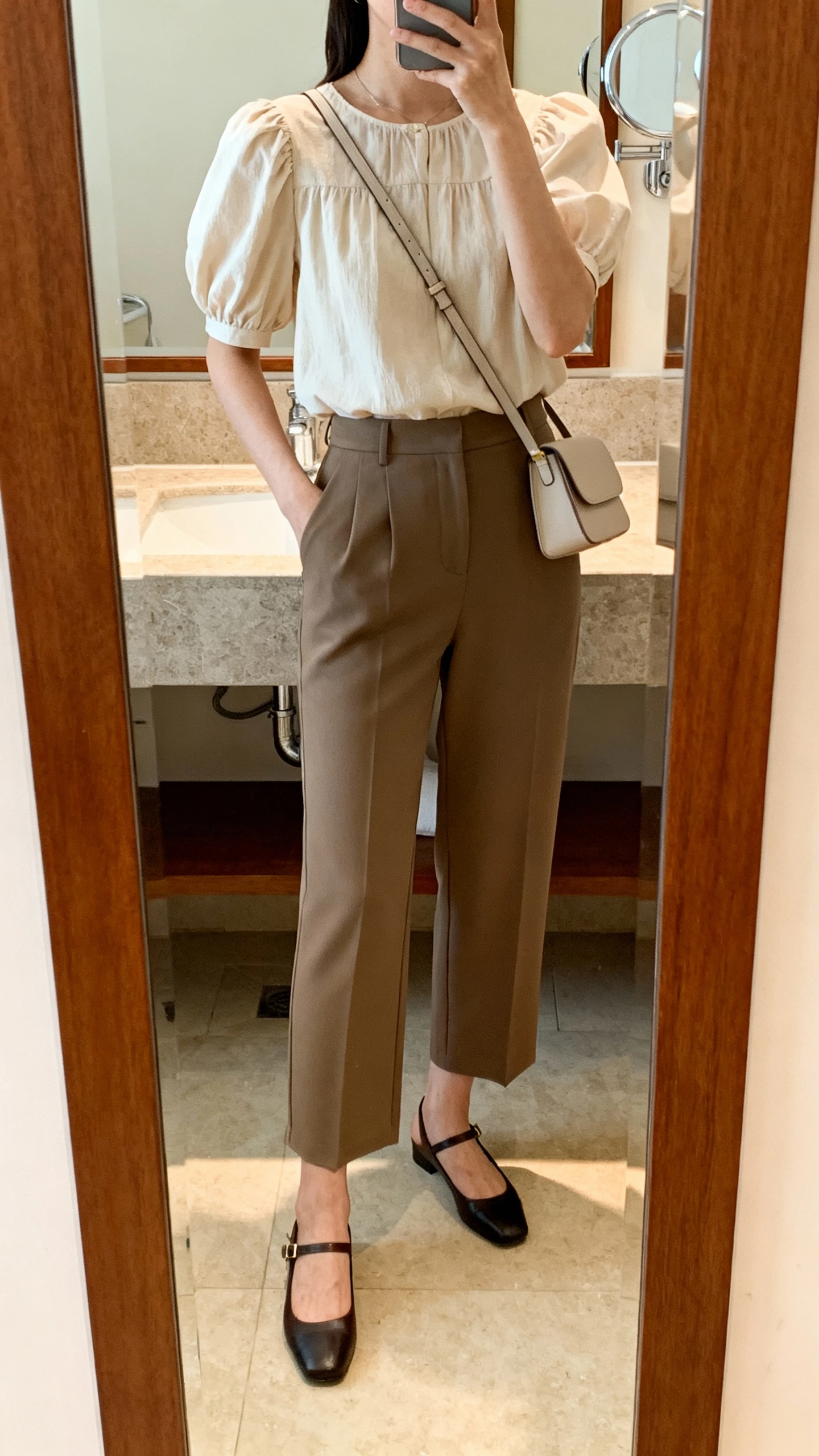 iPhone mirror selfie of a woman wearing a puff-sleeve blouse with tailored culottes, ankle-strap flats, and a minimal crossbody bag, face not visible, cropped at neck, bathroom mirror, warm indoor lighting, iPhone photo quality.