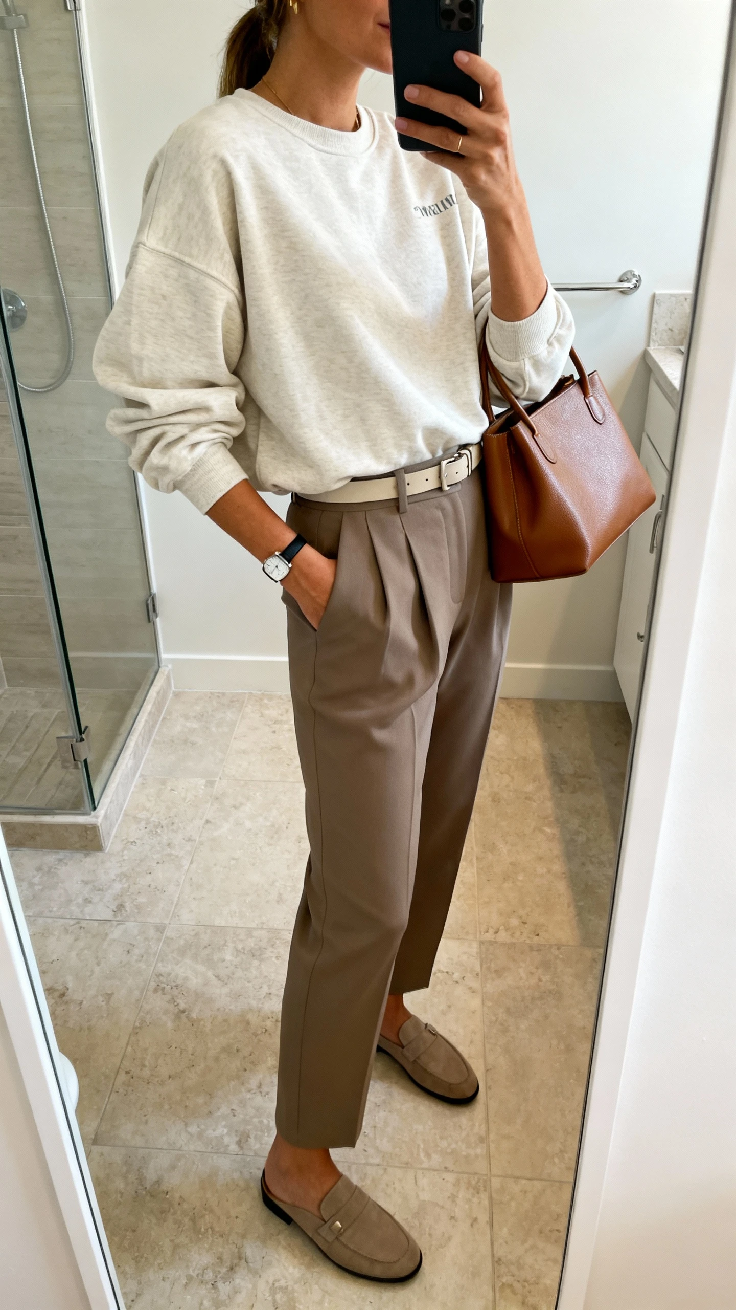 iPhone mirror selfie of a woman wearing a relaxed Christian crewneck with tailored high-waist trousers, minimalist belt, loafers, slim watch, and a small leather tote; face not visible, side angle, bathroom mirror, natural indoor lighting, iPhone photo quality.