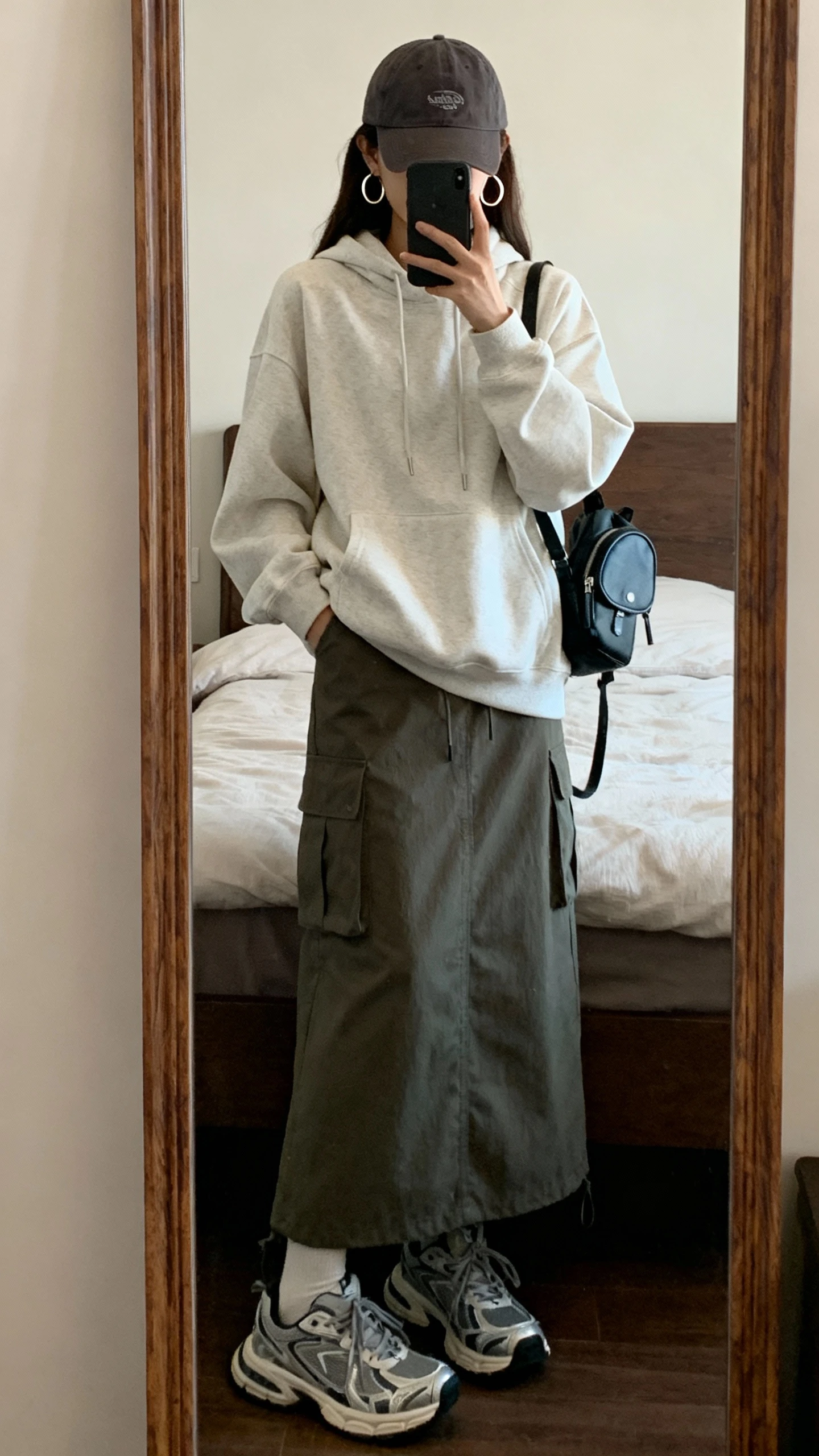 iPhone mirror selfie of a woman wearing a relaxed hoodie with a cargo maxi skirt, chunky trainers, baseball cap, small backpack, hoop earrings, face not visible, cropped at shoulders, bedroom mirror, casual indoor lighting, iPhone photo quality.