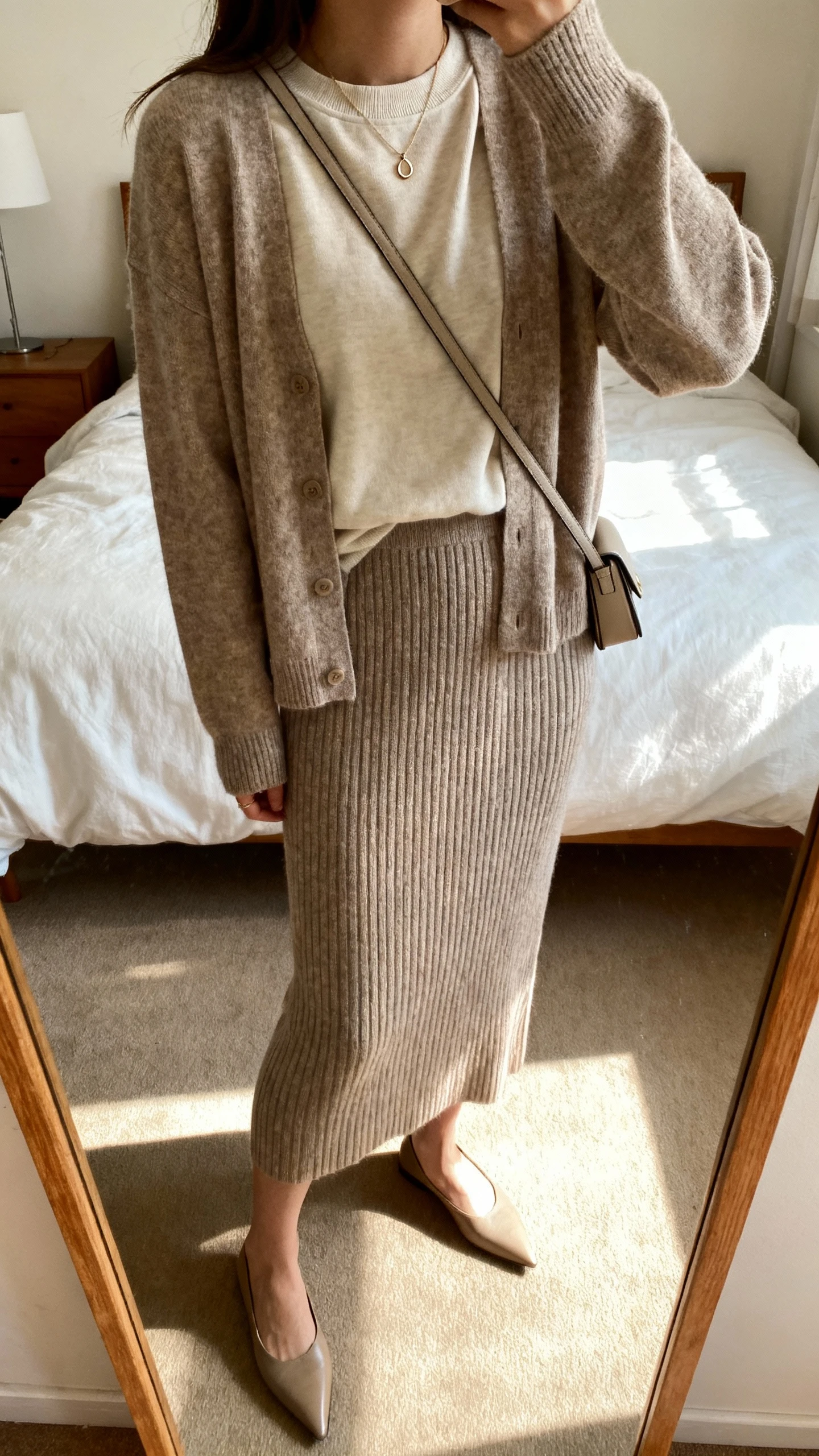 iPhone mirror selfie of a woman wearing a ribbed knit skirt-and-cardigan set with a lightweight crewneck layered underneath, pointed flats, dainty pendant, and a slim crossbody bag; face not visible, cropped at shoulders, bedroom mirror, warm indoor lighting, iPhone photo quality.