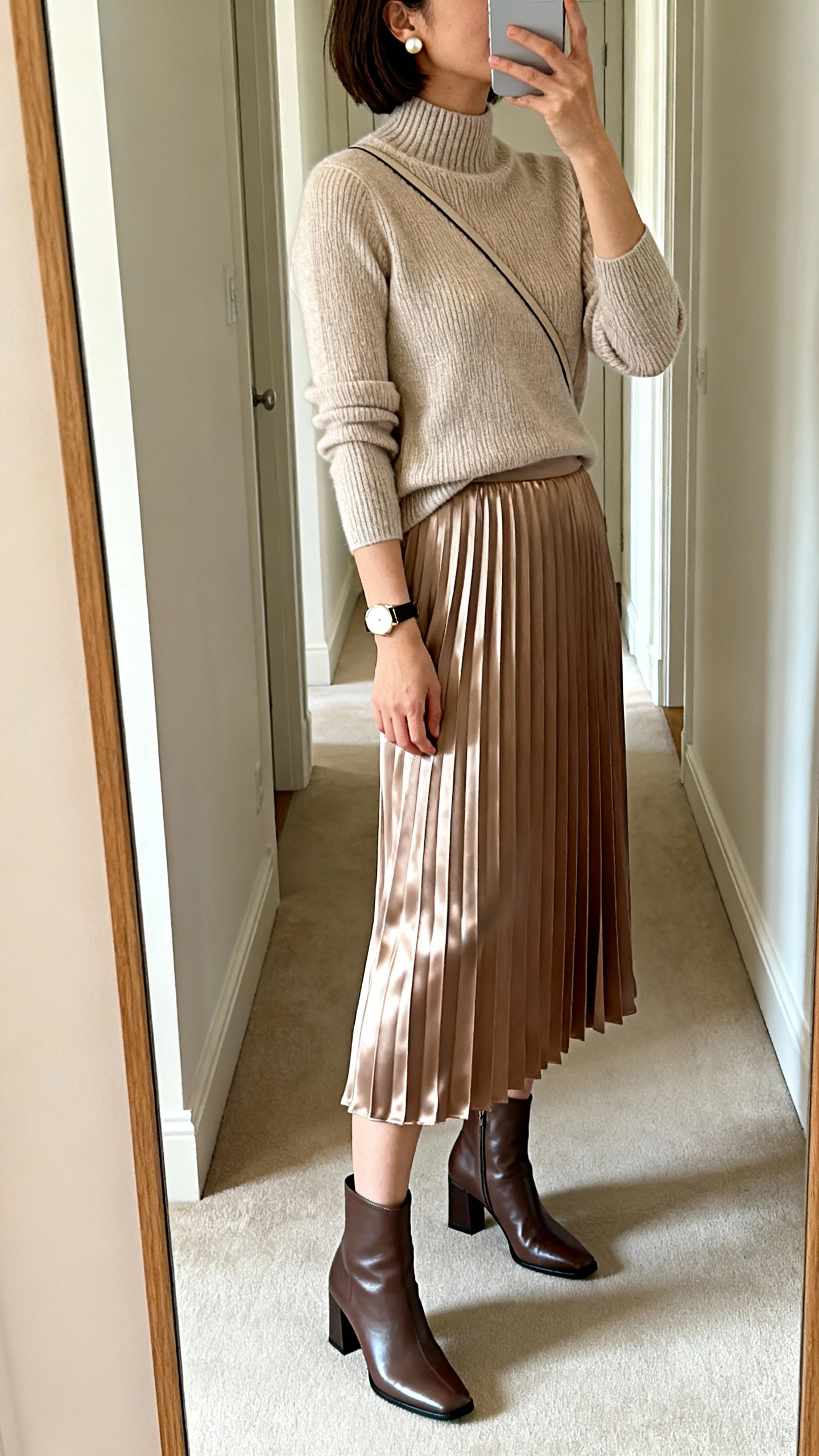 iPhone mirror selfie of a woman wearing a satin pleated midi skirt with a fine-gauge knit mock-neck top, block-heel ankle boots, a simple watch, pearl stud earrings, and a minimal slim crossbody; face not visible, side angle, full-length mirror in hallway, soft morning light, iPhone photo quality.