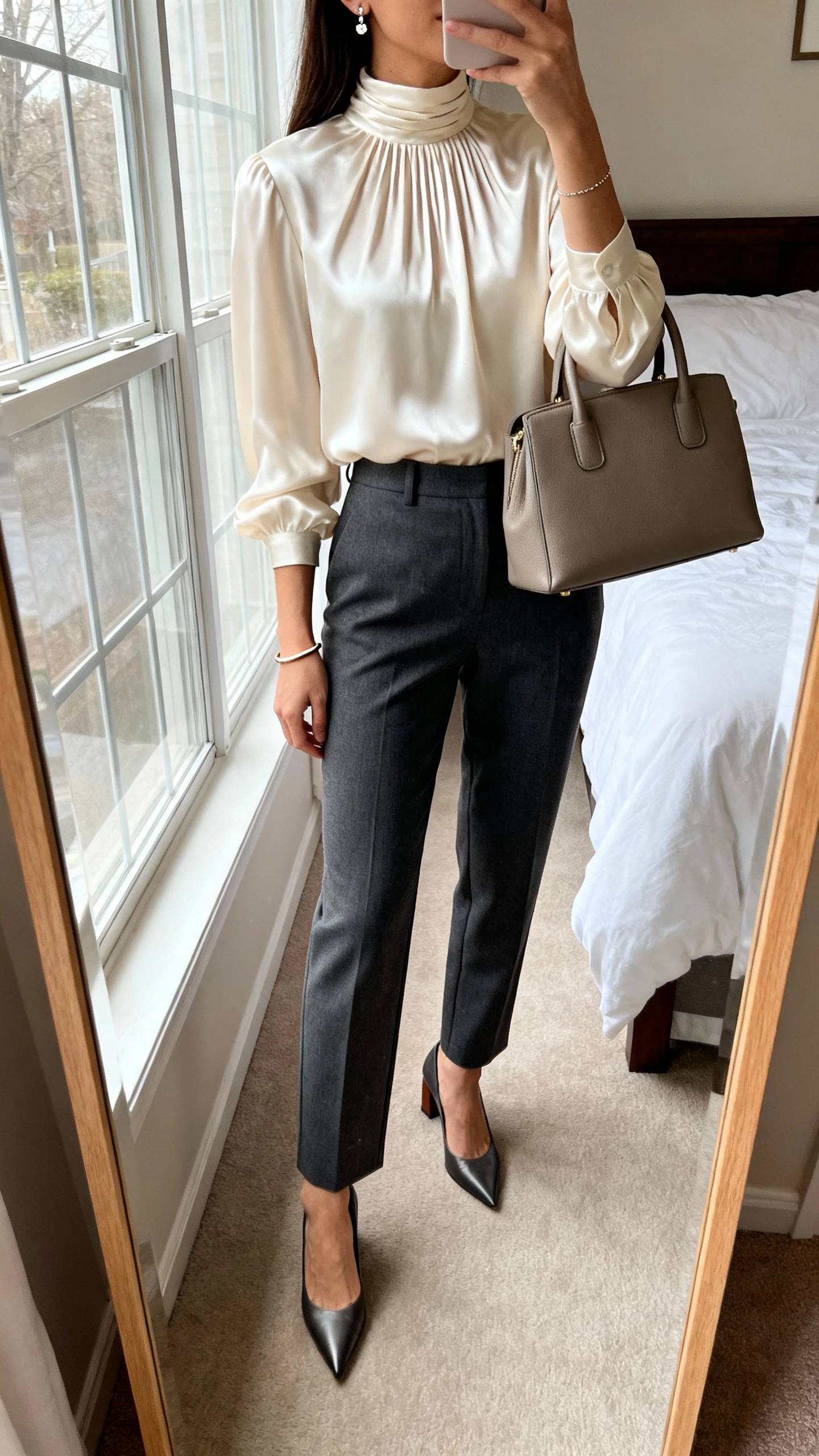 iPhone mirror selfie of a woman wearing a silky high-neck blouse with subtle pleats (cream) tucked into tailored straight-leg trousers (charcoal), pointed-toe block heels, a structured top-handle bag, delicate stud earrings, and a thin bangle; face not visible, cropped at neck, bedroom mirror, morning window light, iPhone photo quality.
