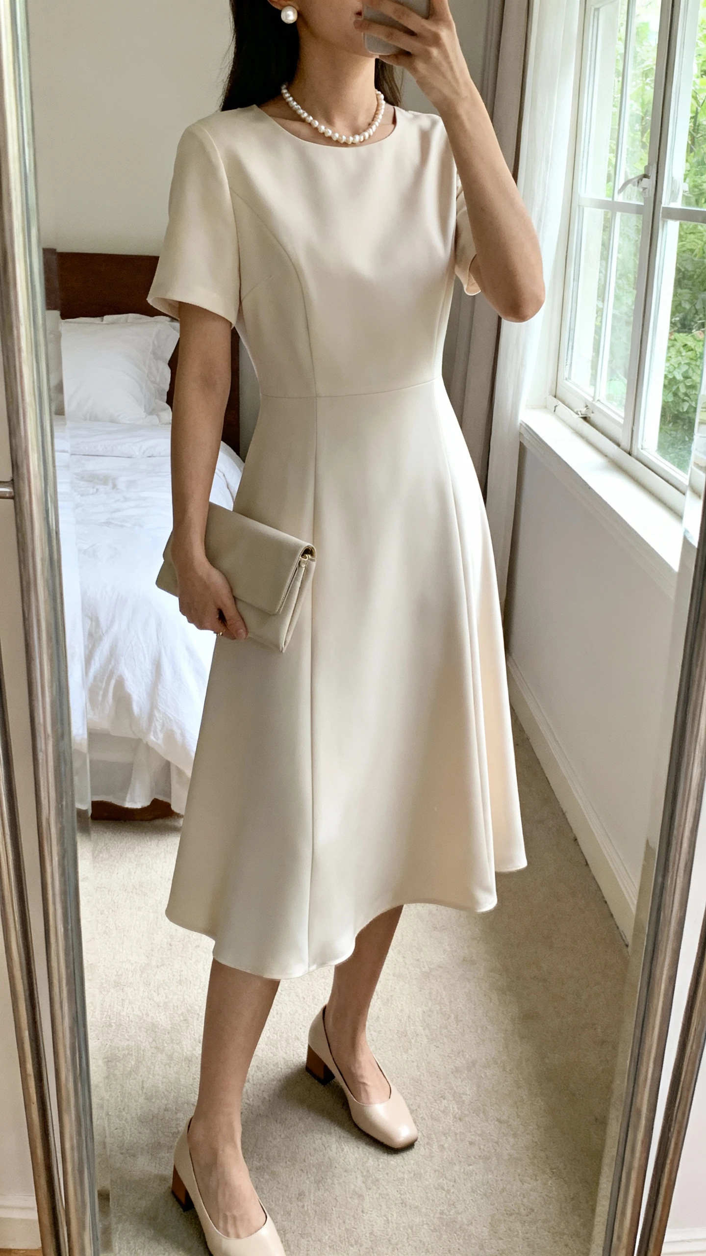 iPhone mirror selfie of a woman wearing a soft A-line midi dress with a polished finish, delicate pearl necklace and pearl stud earrings, simple clutch, and low block heels, face not visible, cropped at neck, bedroom mirror, natural window light, iPhone photo quality.