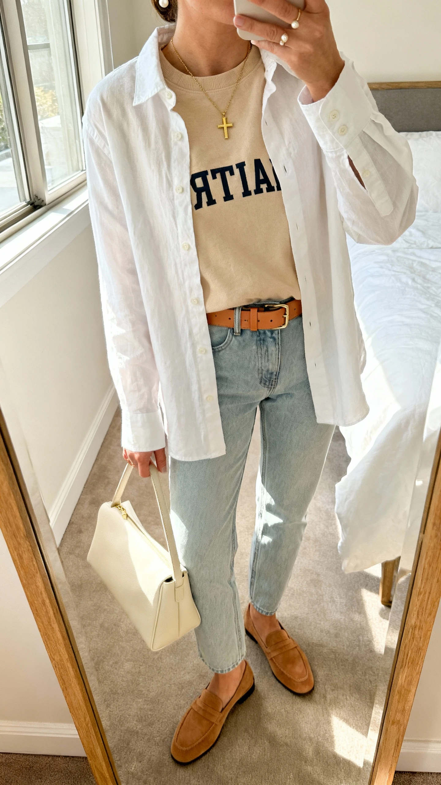 iPhone mirror selfie of a woman wearing a soft beige Christian crewneck layered over a white cotton button-up, light wash straight jeans, tan leather belt, delicate gold cross necklace, small pearl studs, camel loafers, and a cream structured tote, face not visible, cropped at neck, bedroom mirror, morning window light, iPhone photo quality.