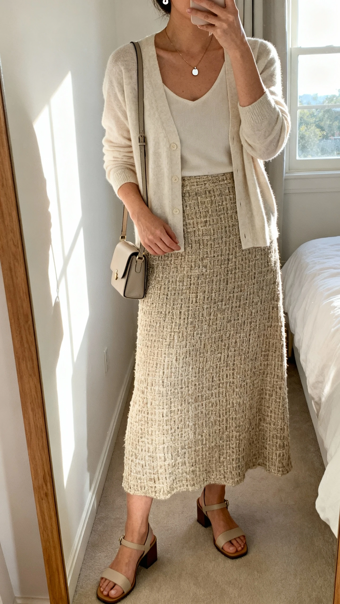 iPhone mirror selfie of a woman wearing a soft cardigan layered over a textured woven-fabric maxi skirt, minimalist pendant necklace, simple stud earrings, low block-heel sandals, and a small crossbody bag; face not visible, cropped at neck, bedroom mirror with natural window light, iPhone photo quality.