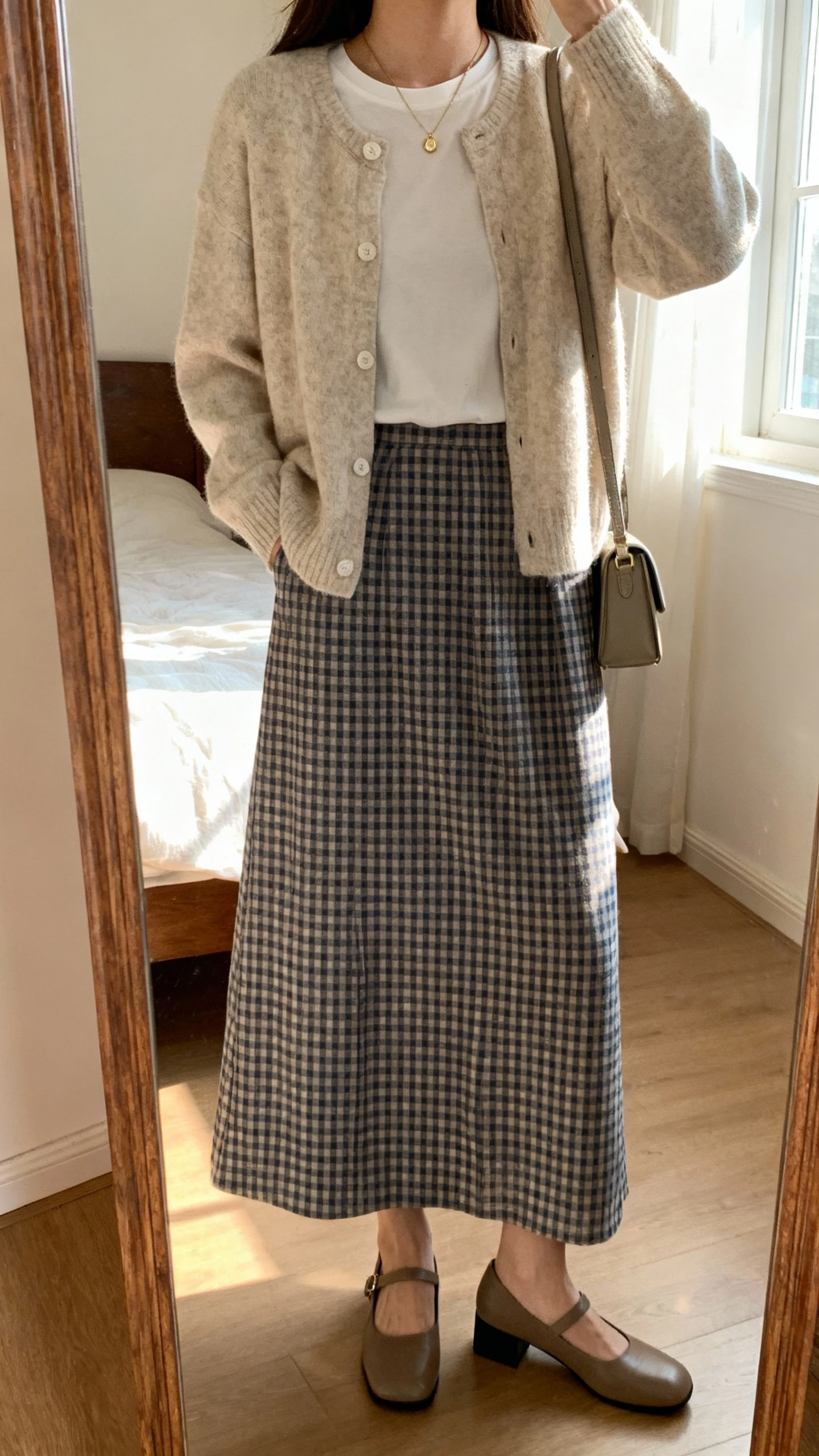 iPhone mirror selfie of a woman wearing a soft cardigan over a simple tucked-in tee, gingham long skirt, low block-heel Mary Janes, delicate pendant necklace, and a small crossbody bag; face not visible, cropped at neck, bedroom mirror, morning window light, iPhone photo quality.