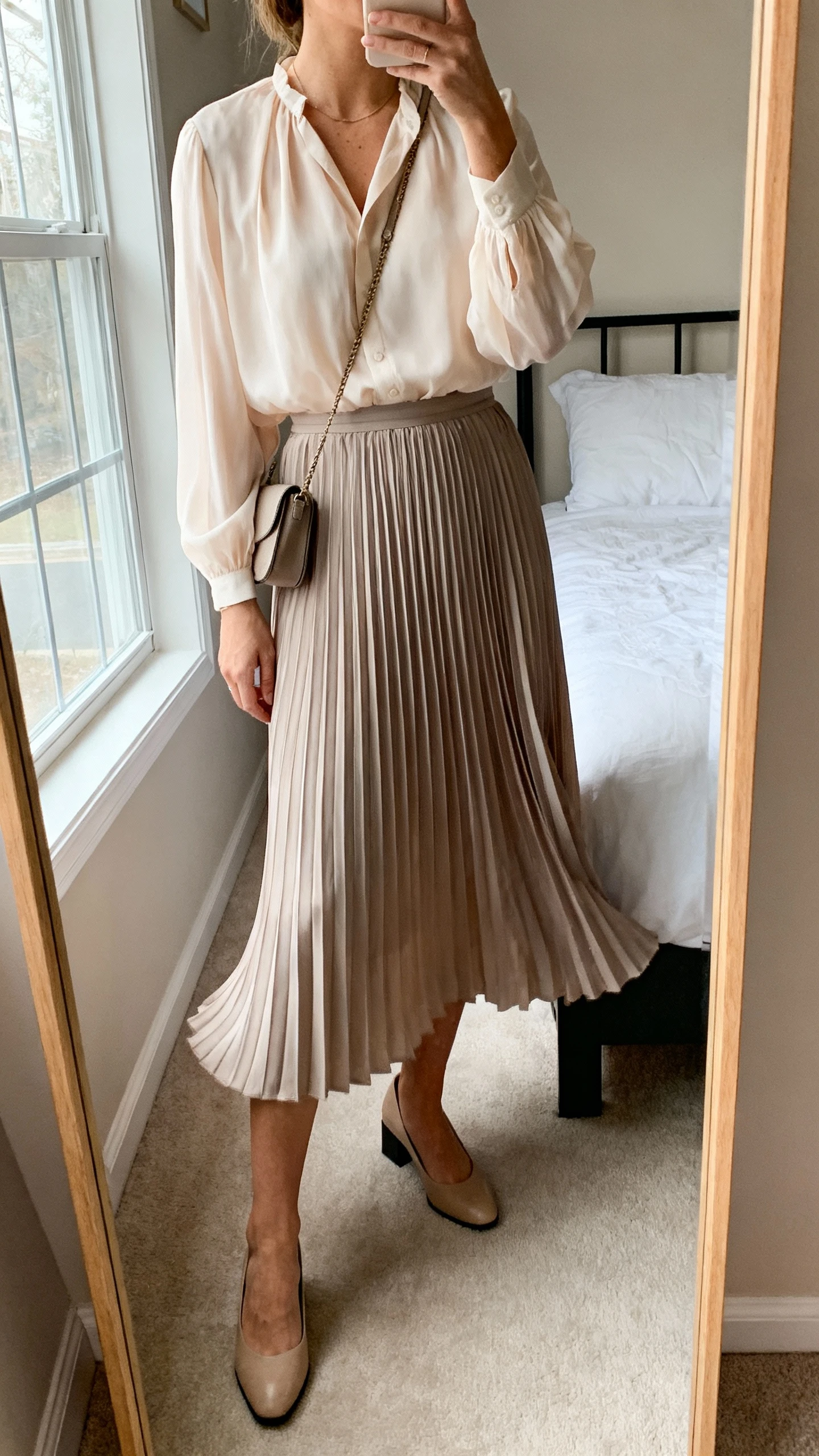 iPhone mirror selfie of a woman wearing a soft chiffon blouse tucked into a pleated midi skirt that sways, paired with low block-heel pumps and a small crossbody, face not visible, cropped at neck, bedroom mirror, natural window light, iPhone photo quality.