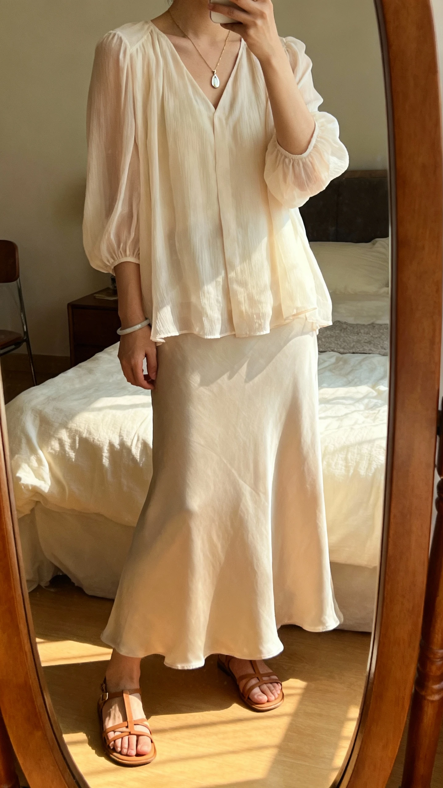 iPhone mirror selfie of a woman wearing a soft chiffon blouse paired with a flowing maxi skirt, styled with a delicate pendant necklace, simple bracelet, and strappy low sandals, face not visible, cropped at shoulders, bedroom mirror with warm afternoon light, iPhone photo quality.