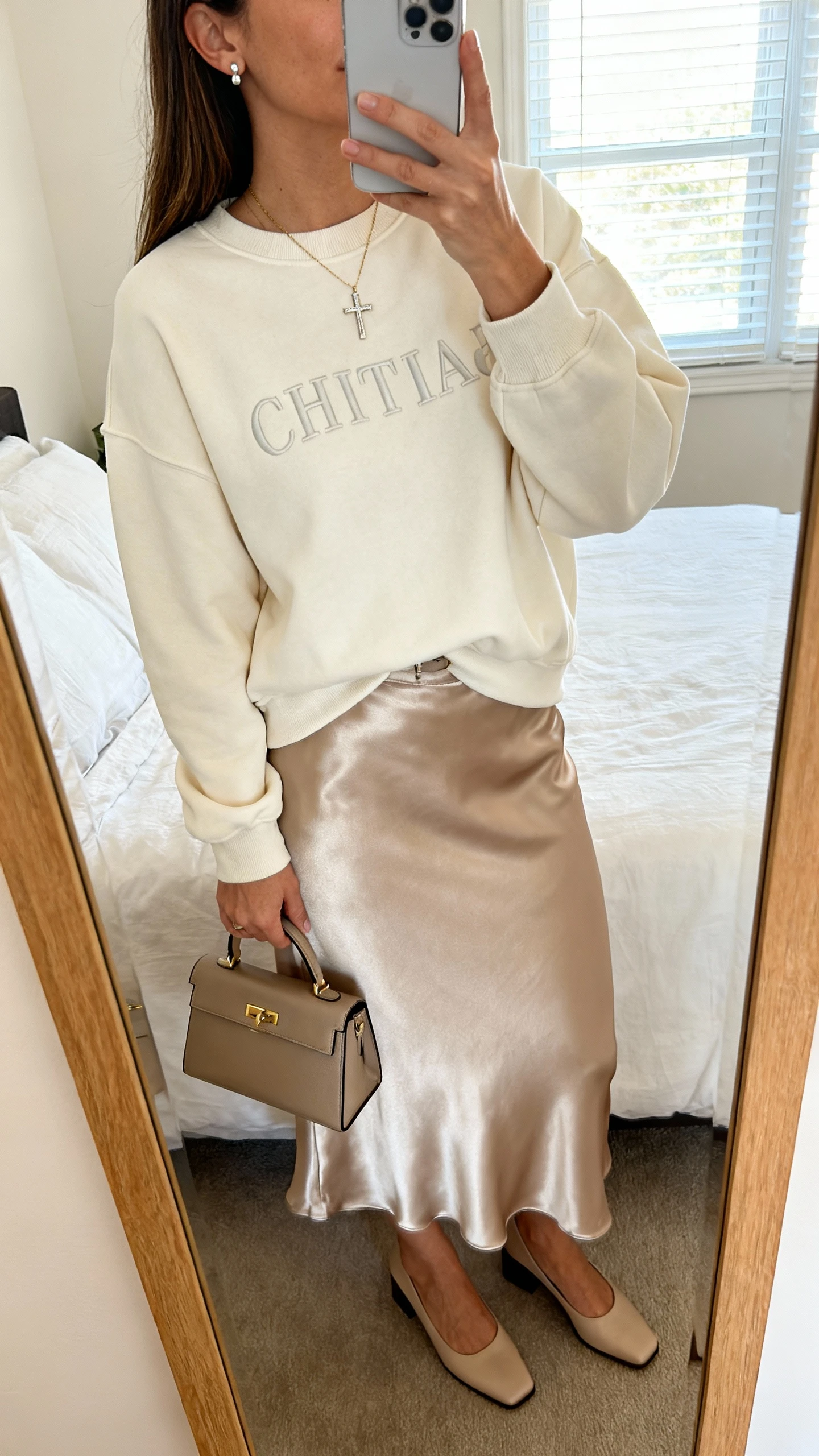 iPhone mirror selfie of a woman wearing a soft Christian crewneck sweatshirt tucked into a satin midi skirt with low block heels, delicate cross necklace, simple stud earrings, and a structured mini handbag; face not visible, cropped at neck, bedroom mirror, morning window light, iPhone photo quality.