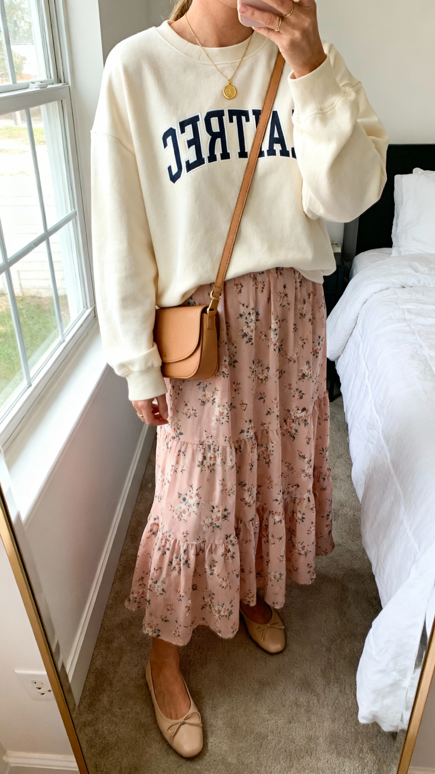 iPhone mirror selfie of a woman wearing a soft cream Christian crewneck layered over a blush floral maxi dress, delicate gold pendant necklace, tan crossbody bag, and beige ballet flats, face not visible, cropped at neck, bedroom mirror, natural morning window light, iPhone photo quality