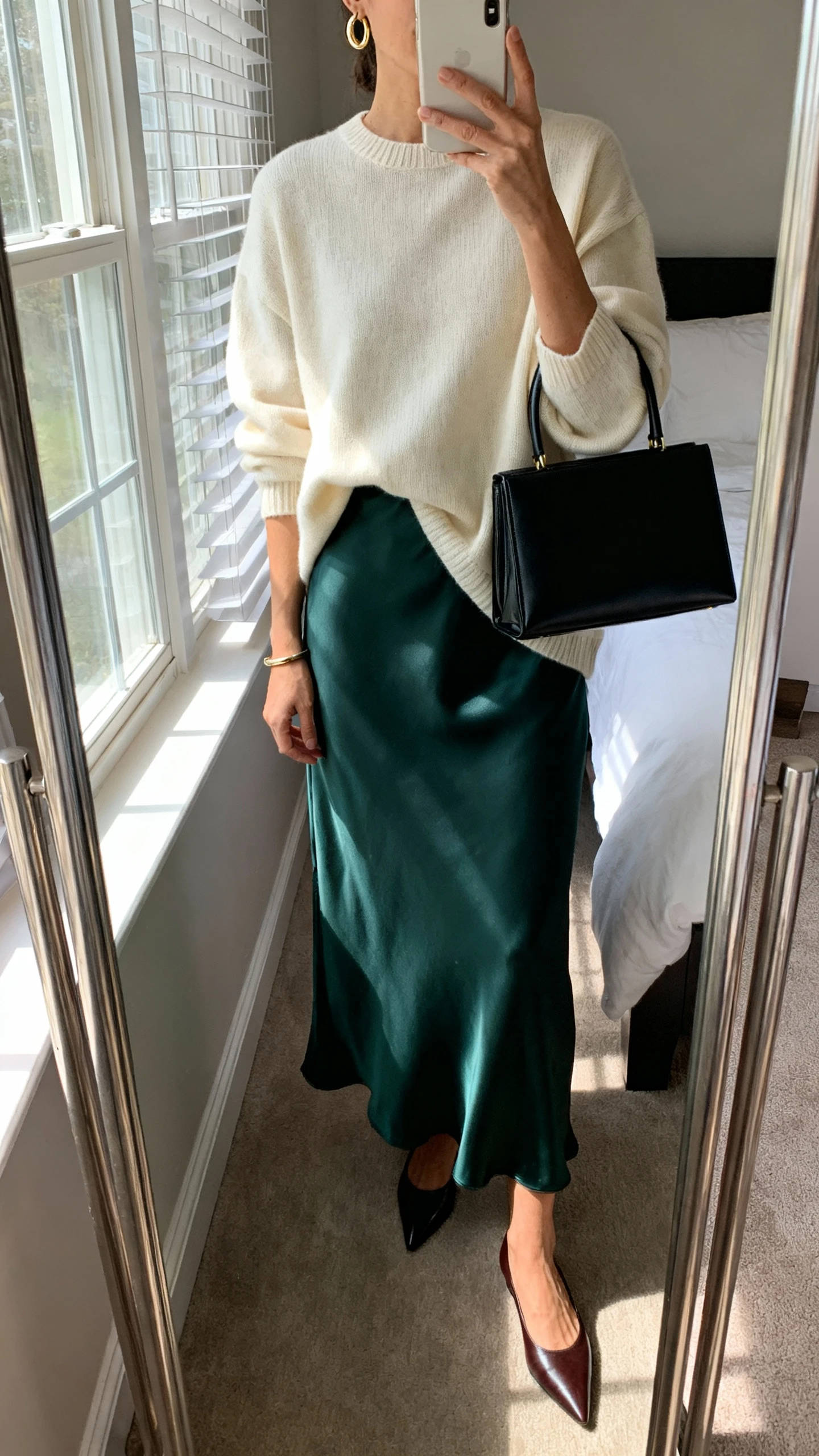 iPhone mirror selfie of a woman wearing a soft cream crewneck sweater (slightly relaxed, hip-length) over a satin maxi dress in deep forest, pointed leather flats, delicate gold hoop earrings, slim bracelet, and a structured top-handle bag in black; face not visible, cropped at neck, bedroom mirror with natural window light, iPhone photo quality.