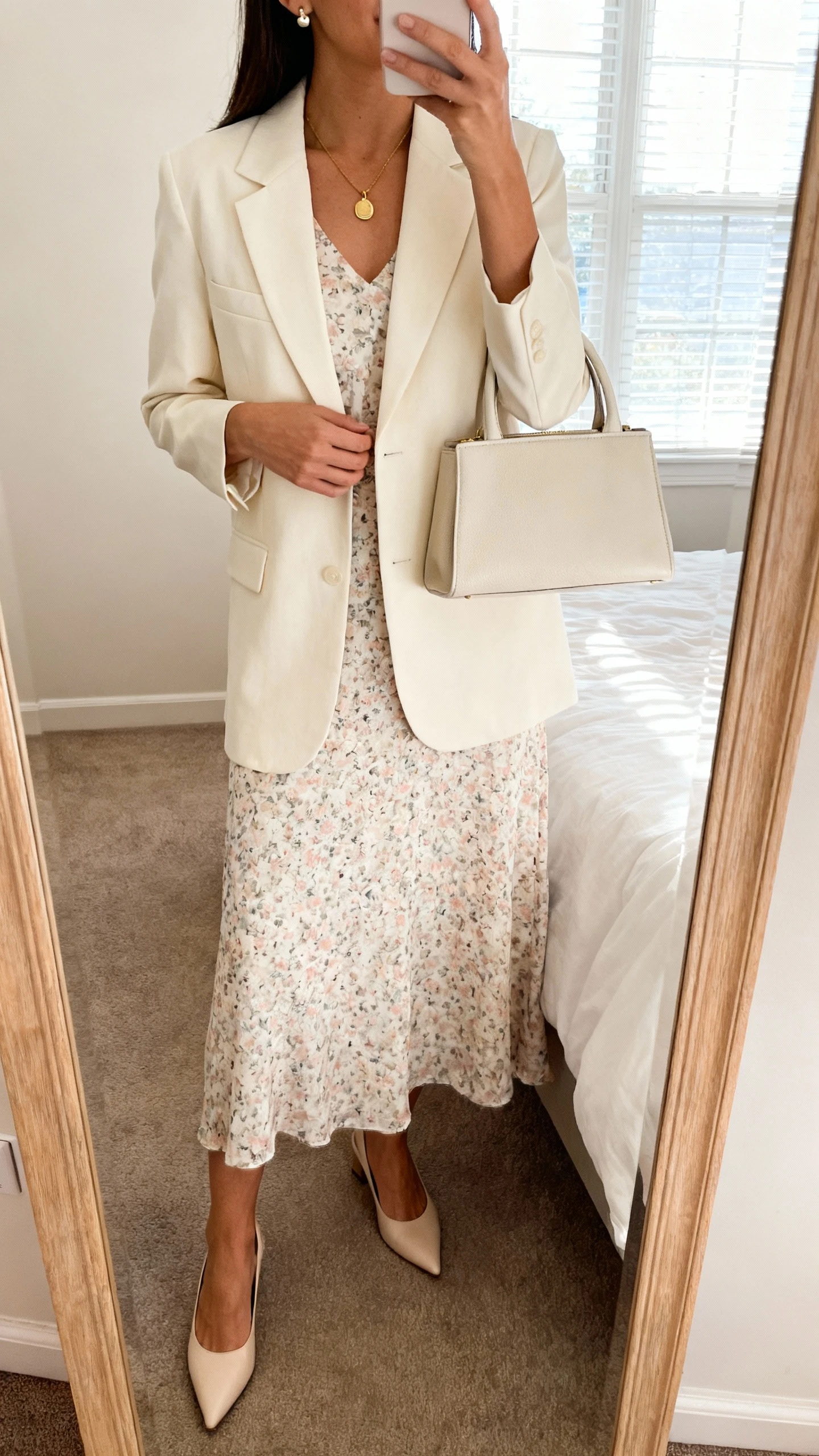 iPhone mirror selfie of a woman wearing a soft floral midi dress, a cream tailored blazer, pointed-toe low heels, a simple gold pendant necklace with stud earrings, and a structured top-handle bag, face not visible, cropped at neck, bedroom mirror, natural window light, iPhone photo quality.