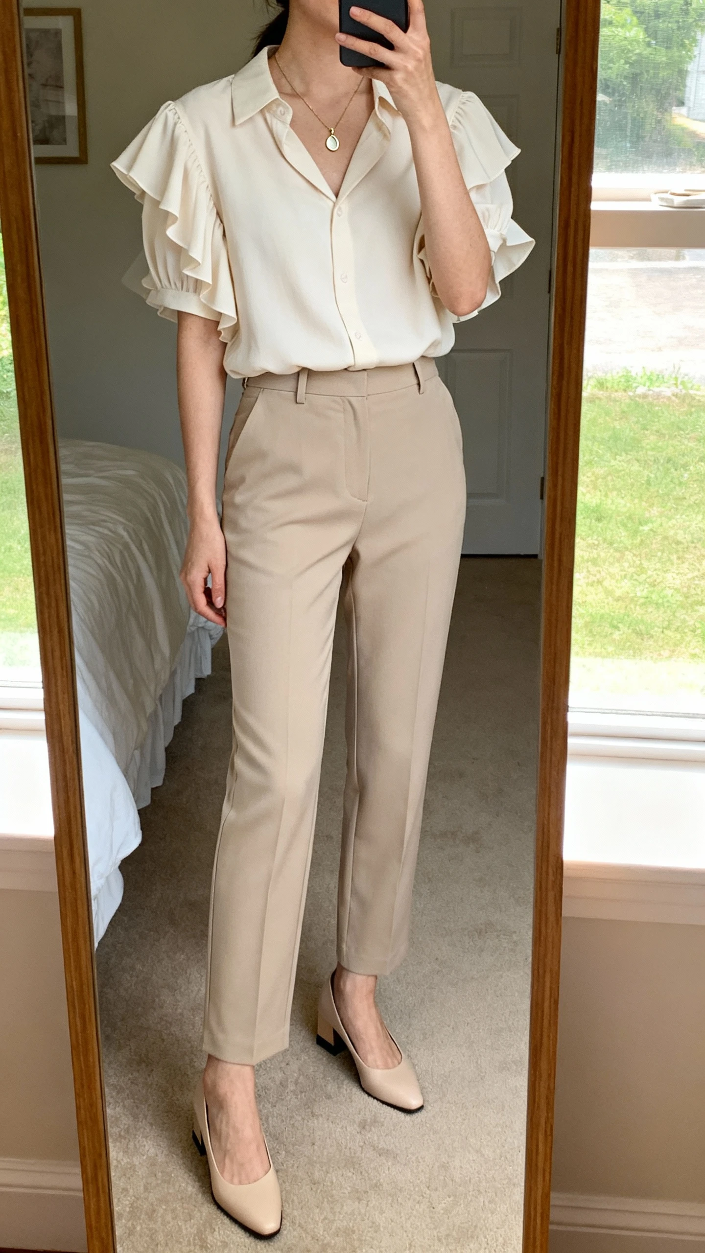 iPhone mirror selfie of a woman wearing a soft flutter-sleeve blouse with a slightly tailored fit tucked into high-waisted straight trousers, delicate pendant necklace, low block-heel pumps, face not visible, cropped at neck, bedroom mirror, natural window light, iPhone photo quality.