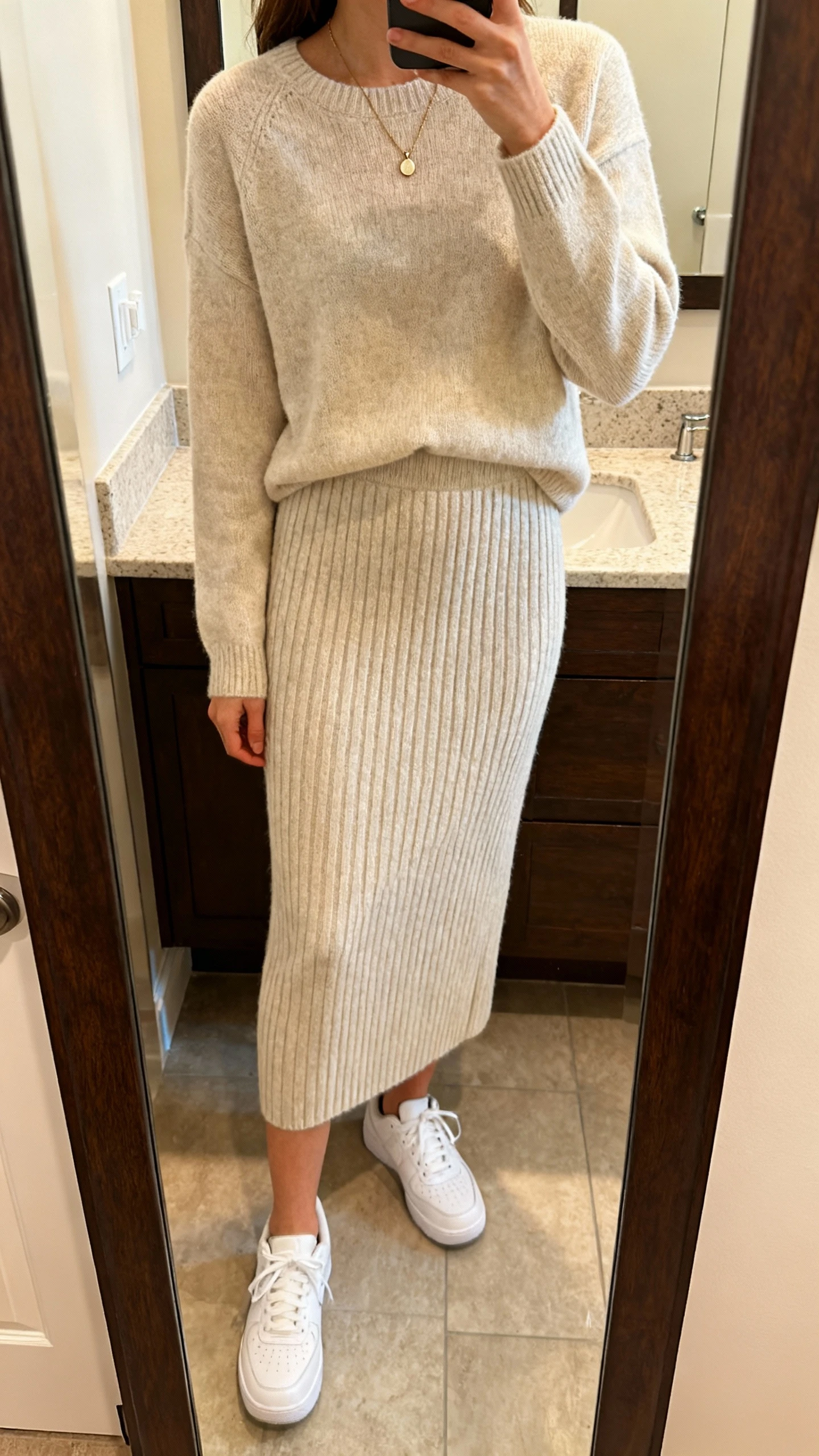 iPhone mirror selfie of a woman wearing a soft knit matching skirt set (ribbed midi skirt with coordinating knit top), white sneakers, delicate pendant necklace, face not visible, bathroom full-length mirror, warm indoor lighting, iPhone photo quality.