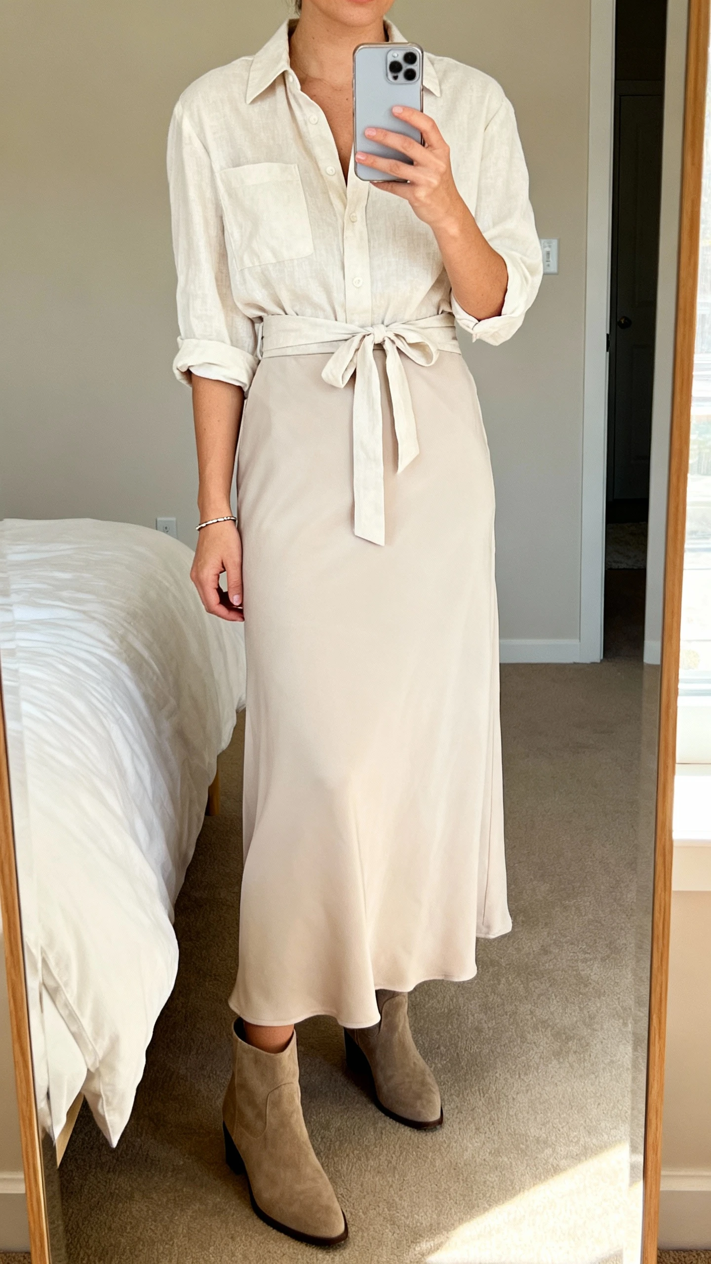 iPhone mirror selfie of a woman wearing a soft maxi skirt, a belted blouse, ankle boots, and a subtle bracelet; face not visible, cropped at neck, bedroom mirror with warm indoor lighting, casual iPhone photo.