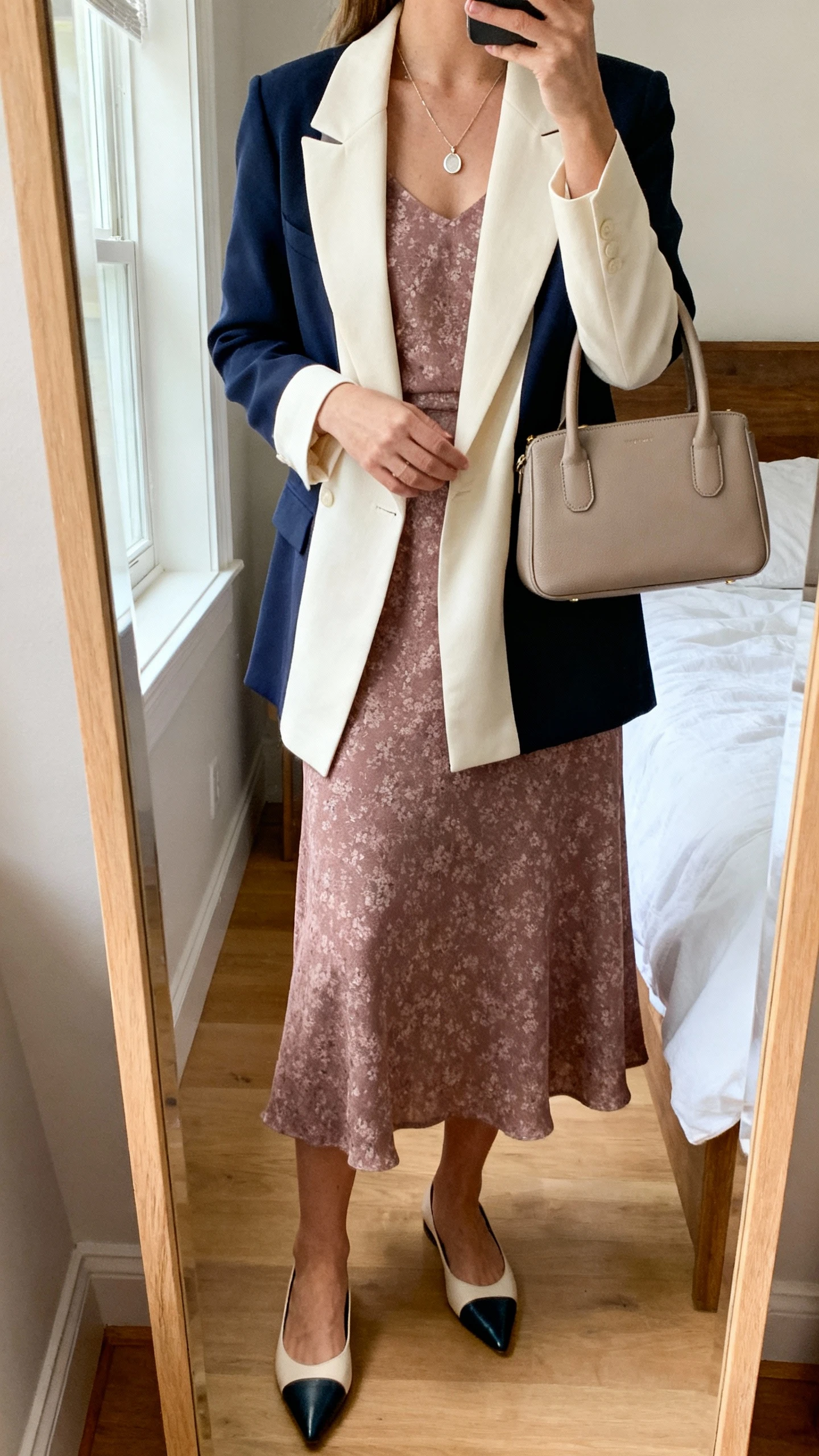 iPhone mirror selfie of a woman wearing a soft midi dress in a subtle floral or jewel tone with a tailored cream or navy blazer, pointed-toe flats, a delicate pendant necklace, and a structured neutral top-handle bag; face not visible, cropped at neck, bedroom mirror, natural window light, iPhone photo quality.