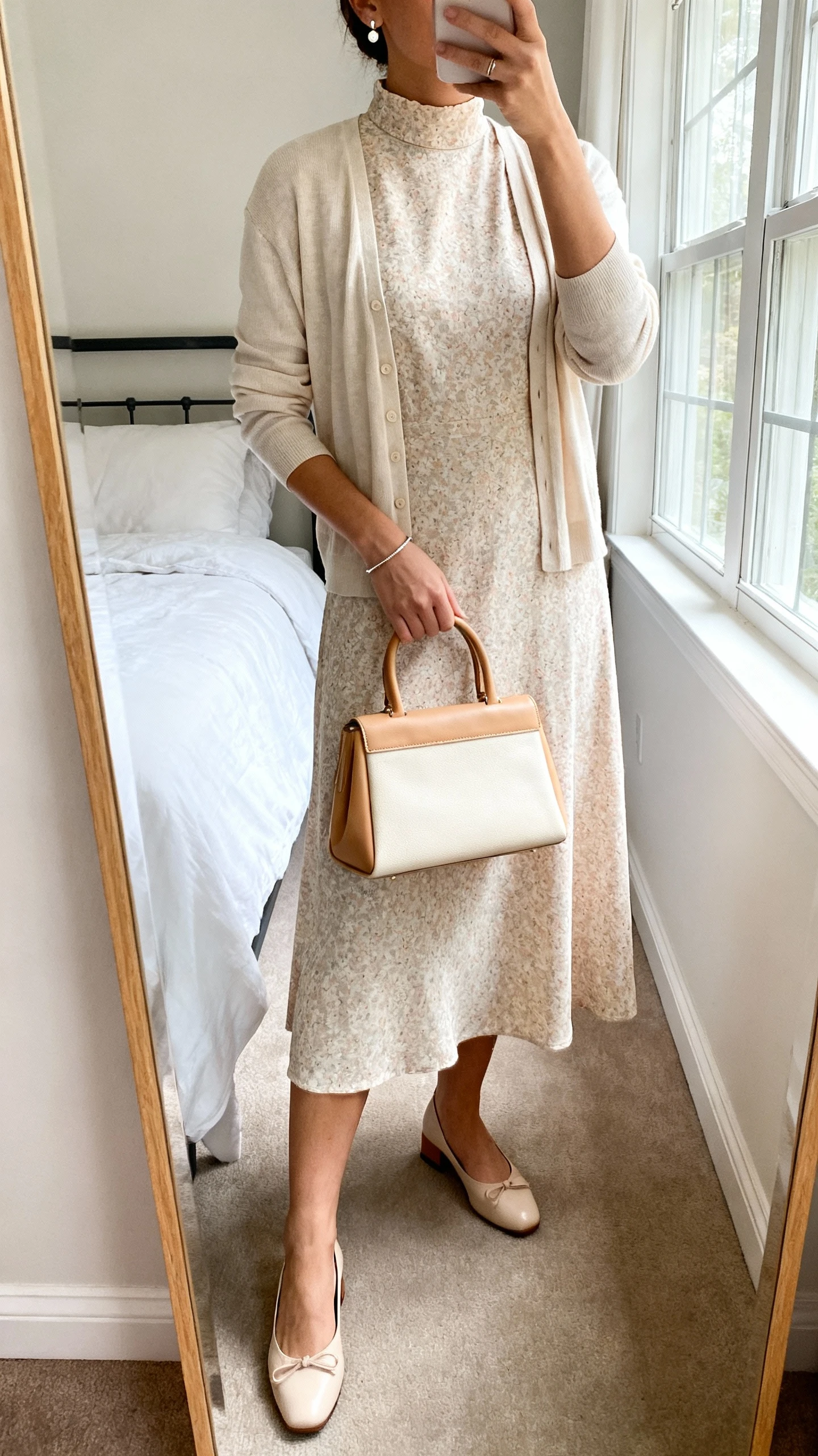 iPhone mirror selfie of a woman wearing a soft midi dress with a high neckline and elbow-length sleeves (floral or solid), a lightweight cardigan, low block heels or classic ballet flats, a structured top-handle bag in tan or cream, delicate stud earrings, and a thin bracelet; face not visible, cropped at neck, bedroom mirror, natural window light, iPhone photo quality.