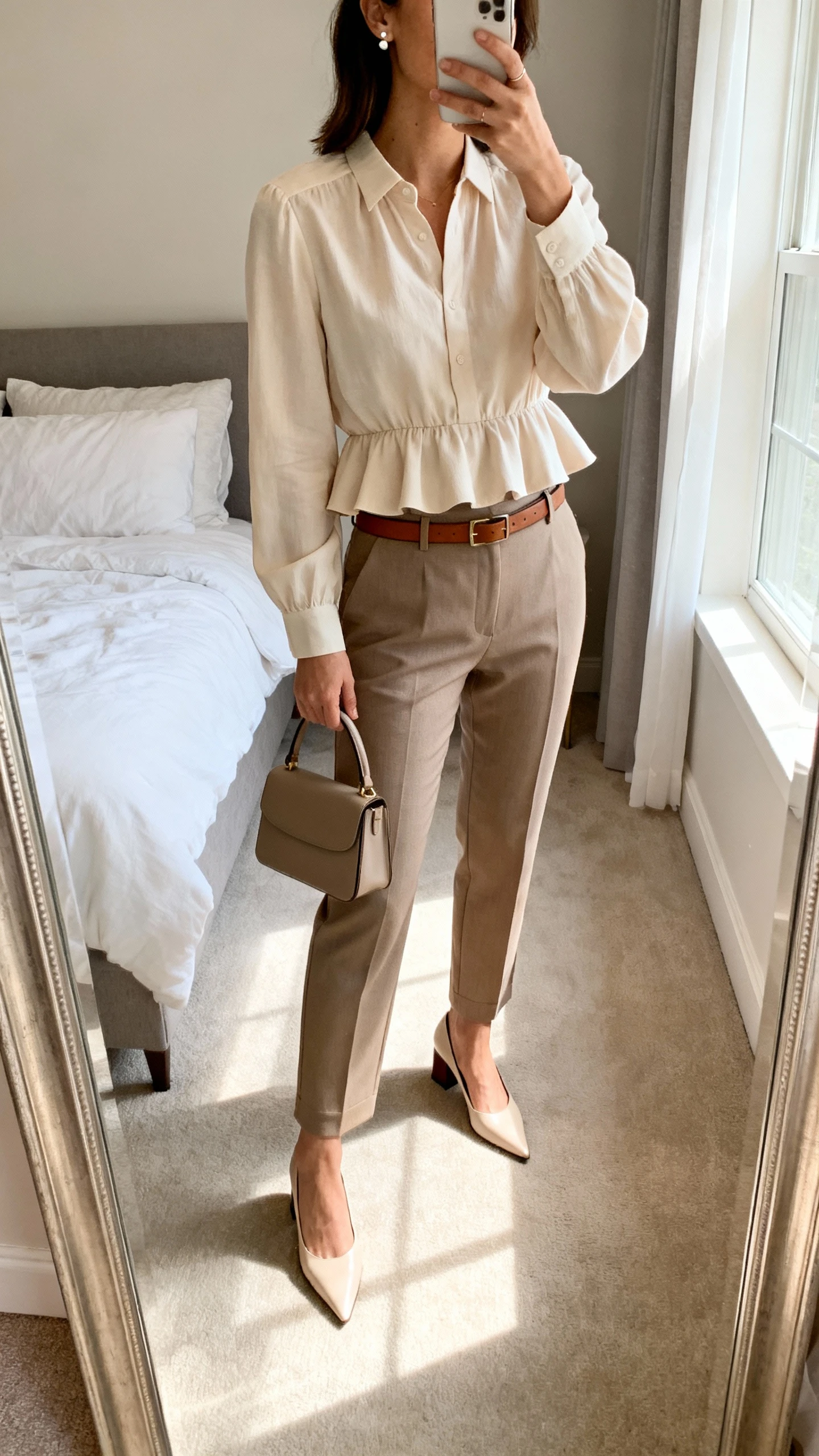 iPhone mirror selfie of a woman wearing a soft-peplum blouse tucked into high-waist tailored trousers with a slim leather belt, pointed-toe block heels, structured top-handle bag, and delicate stud earrings, face not visible, cropped at neck, bedroom mirror, morning window light, iPhone photo quality.