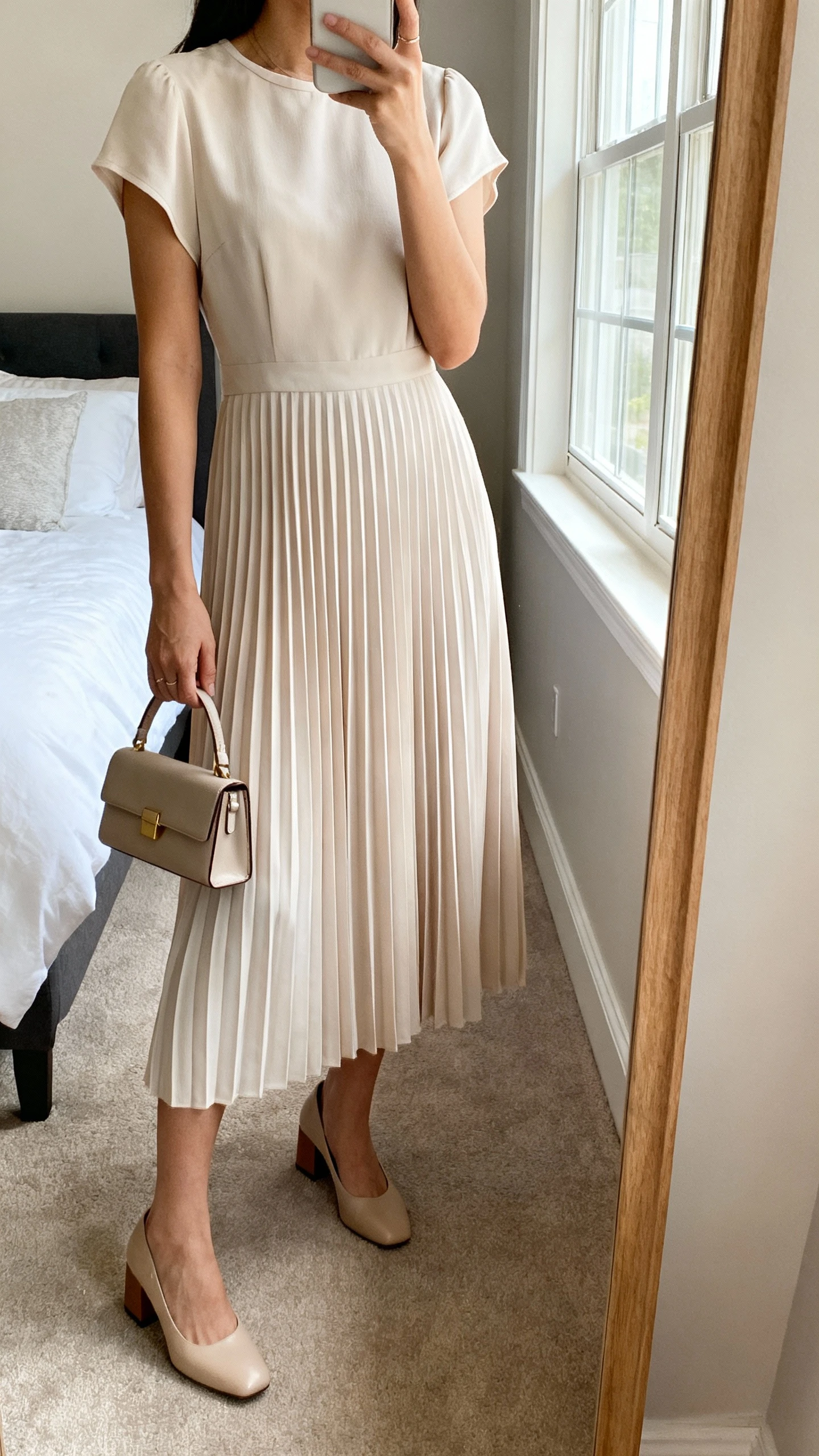 iPhone mirror selfie of a woman wearing a soft-pleat midi dress with a modest neckline and short sleeves, paired with low block-heel pumps and a small structured handbag; face not visible, cropped at neck, bedroom mirror, natural window light, iPhone photo quality.