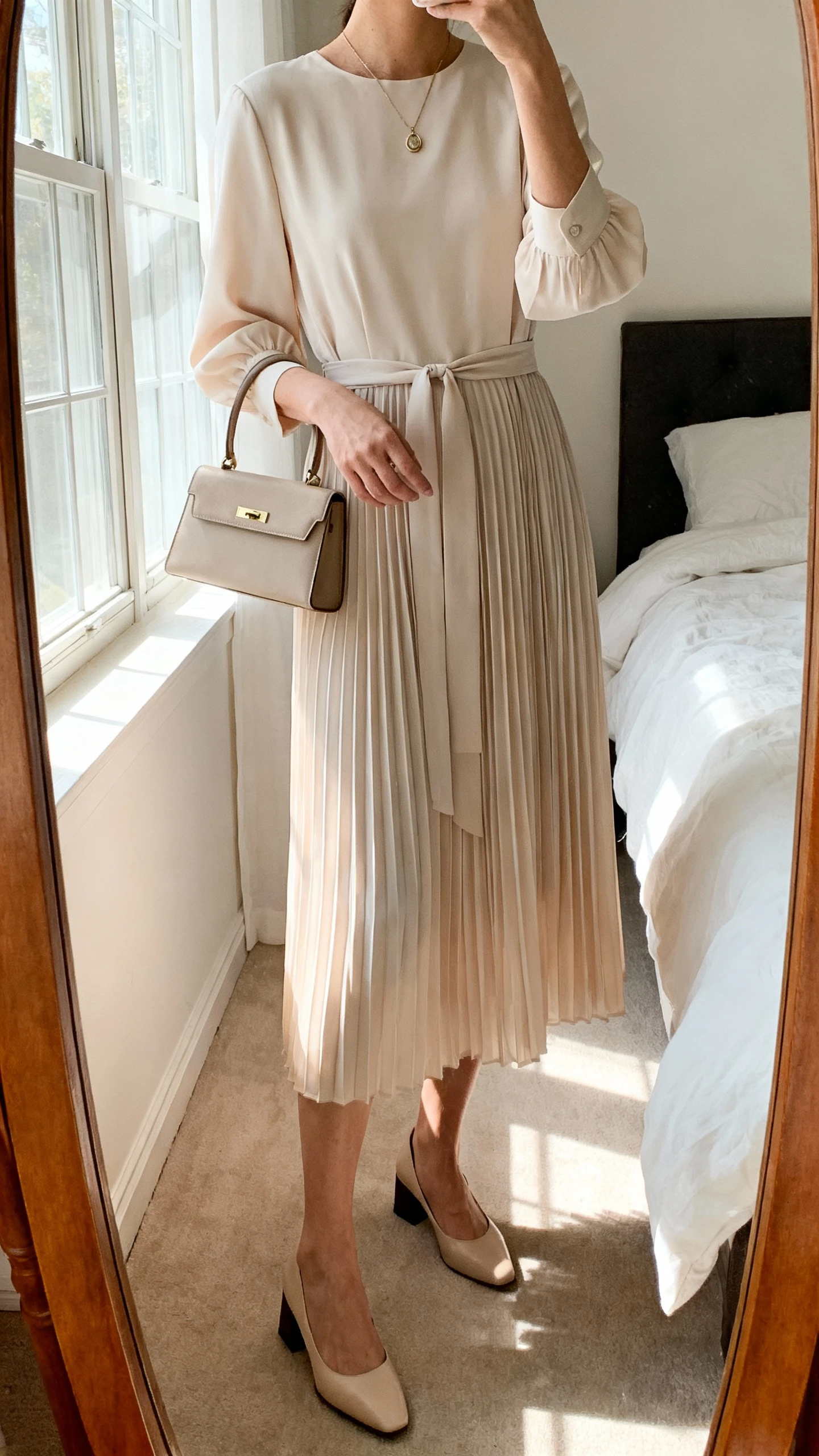 iPhone mirror selfie of a woman wearing a soft-pleat midi dress with modest neckline and elbow sleeves, cinched waist, simple pendant necklace, low block-heel pumps, small structured top-handle bag, face not visible, cropped at neck, bedroom mirror, natural morning window light, iPhone photo quality
