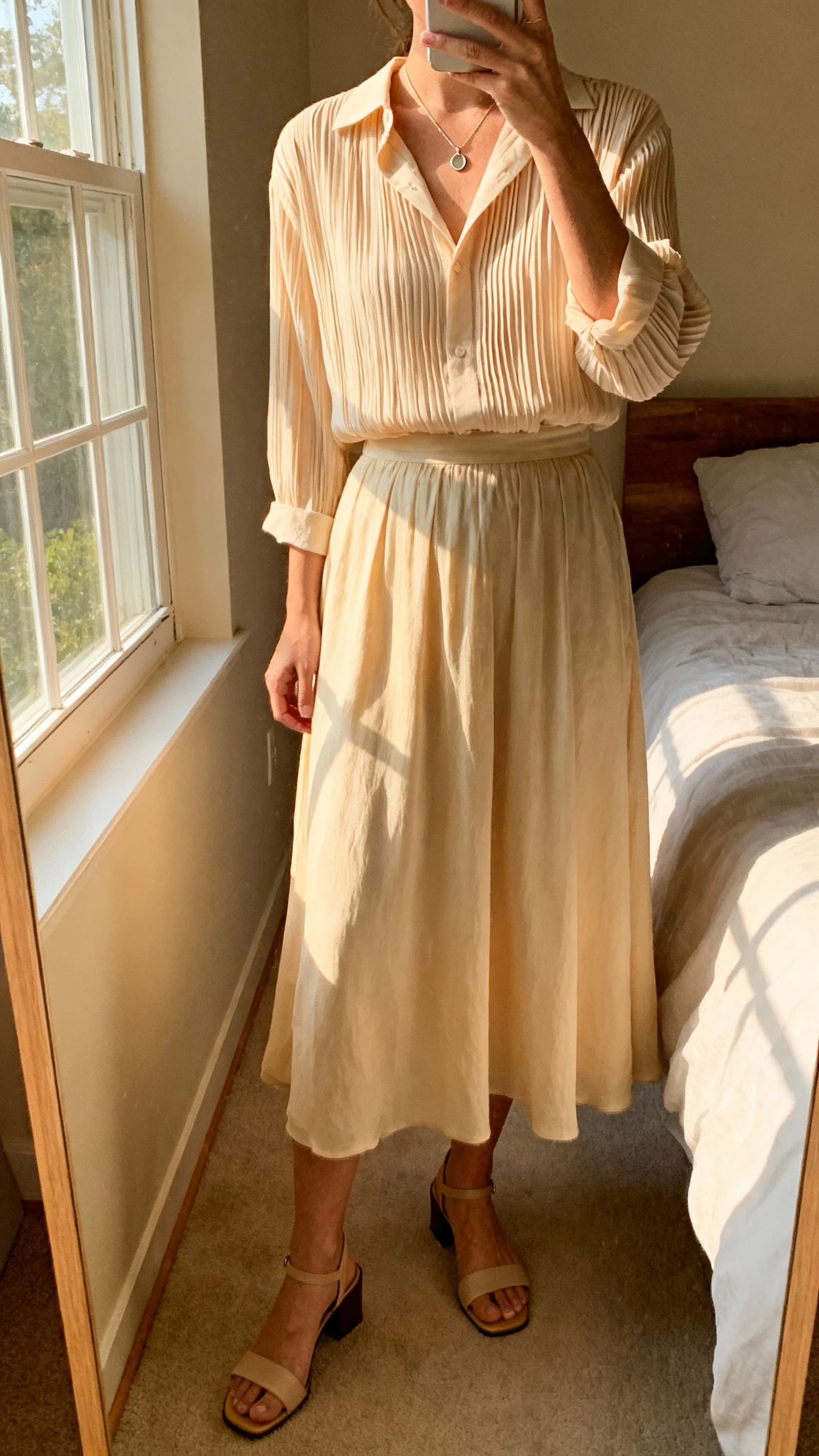 iPhone mirror selfie of a woman wearing a soft-pleated blouse tucked into a flowy midi skirt with low block-heel sandals and a delicate pendant necklace, face not visible, cropped at neck, bedroom mirror with natural window light, iPhone photo quality.