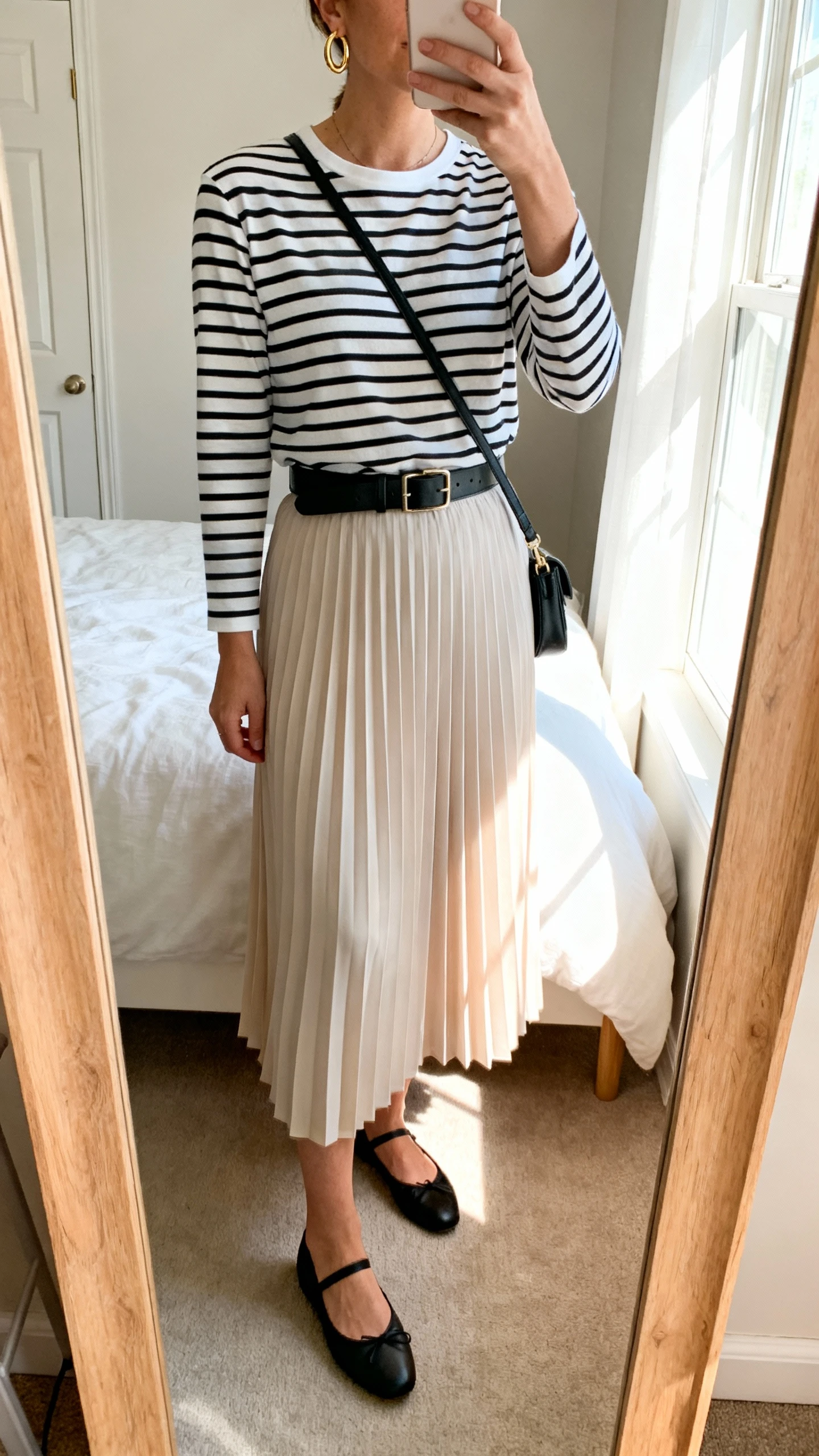 iPhone mirror selfie of a woman wearing a soft-pleated midi skirt, classic black-and-white Breton striped long-sleeve tee, simple black ballet flats, a slim black leather belt, delicate gold hoop earrings, and a small crossbody bag, face not visible, cropped at neck, bedroom mirror with morning window light, iPhone photo quality.