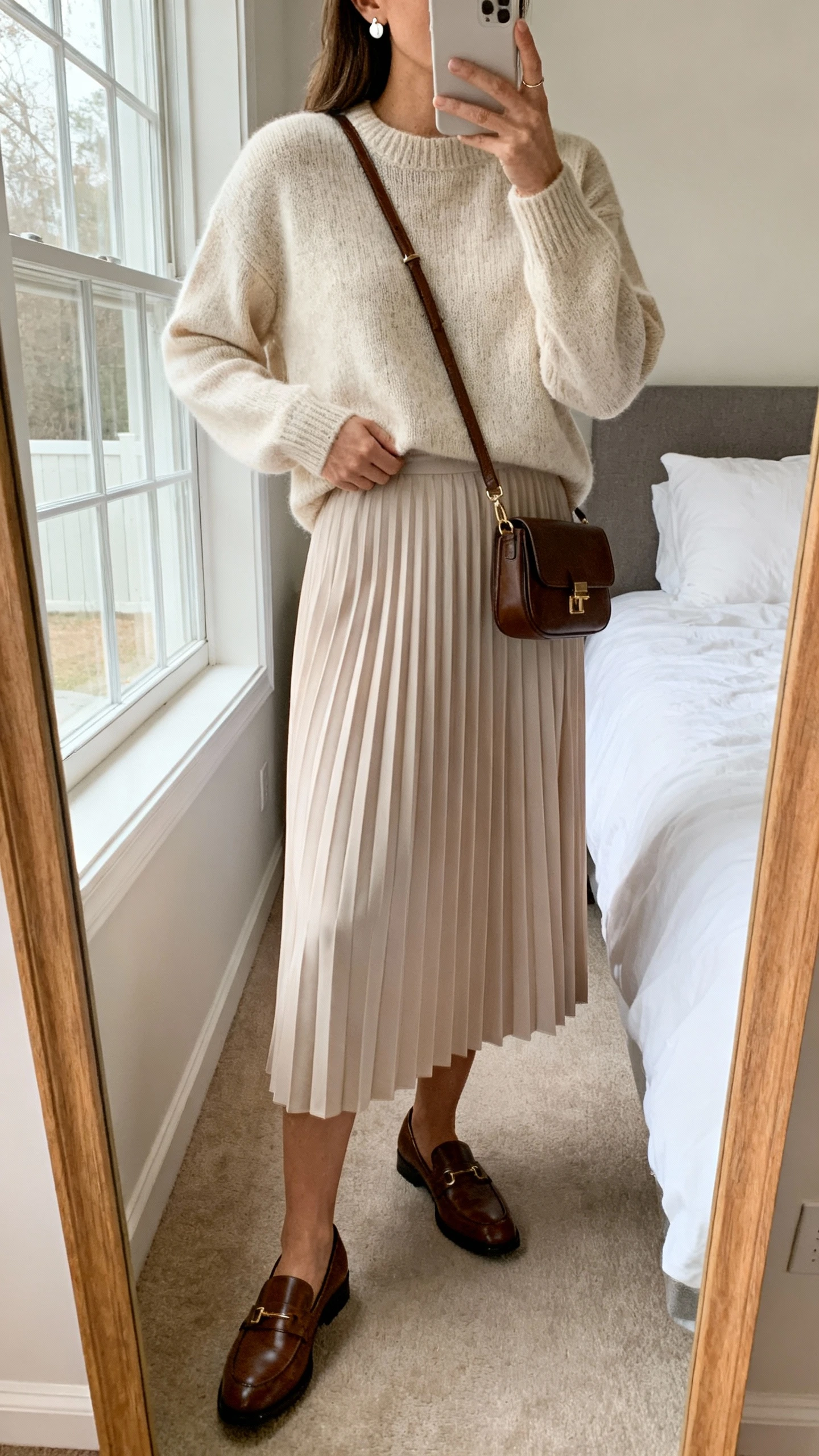 iPhone mirror selfie of a woman wearing a soft pleated midi skirt with a polished knit sweater, simple stud earrings, classic leather loafers, and a small structured crossbody, face not visible, cropped at neck, bedroom mirror with natural window light, iPhone photo quality.