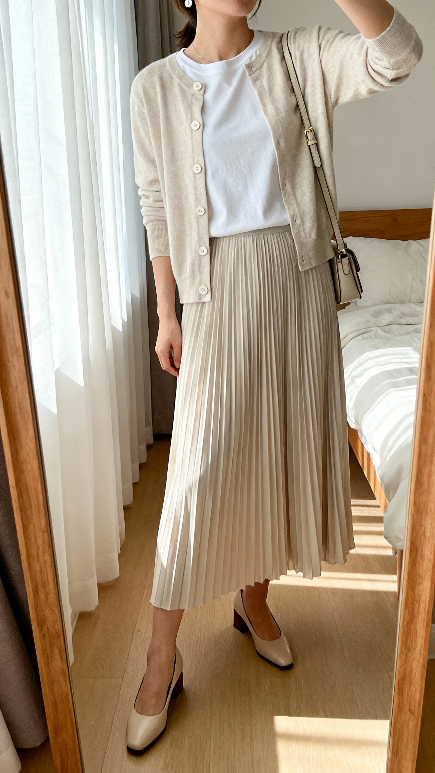 iPhone mirror selfie of a woman wearing a soft-pleated long midi skirt, lightweight buttoned cardigan layered over a simple crewneck tee, low block-heel pumps, delicate stud earrings, and a small crossbody bag, face not visible, cropped at neck, bedroom mirror with morning window light, iPhone photo quality.