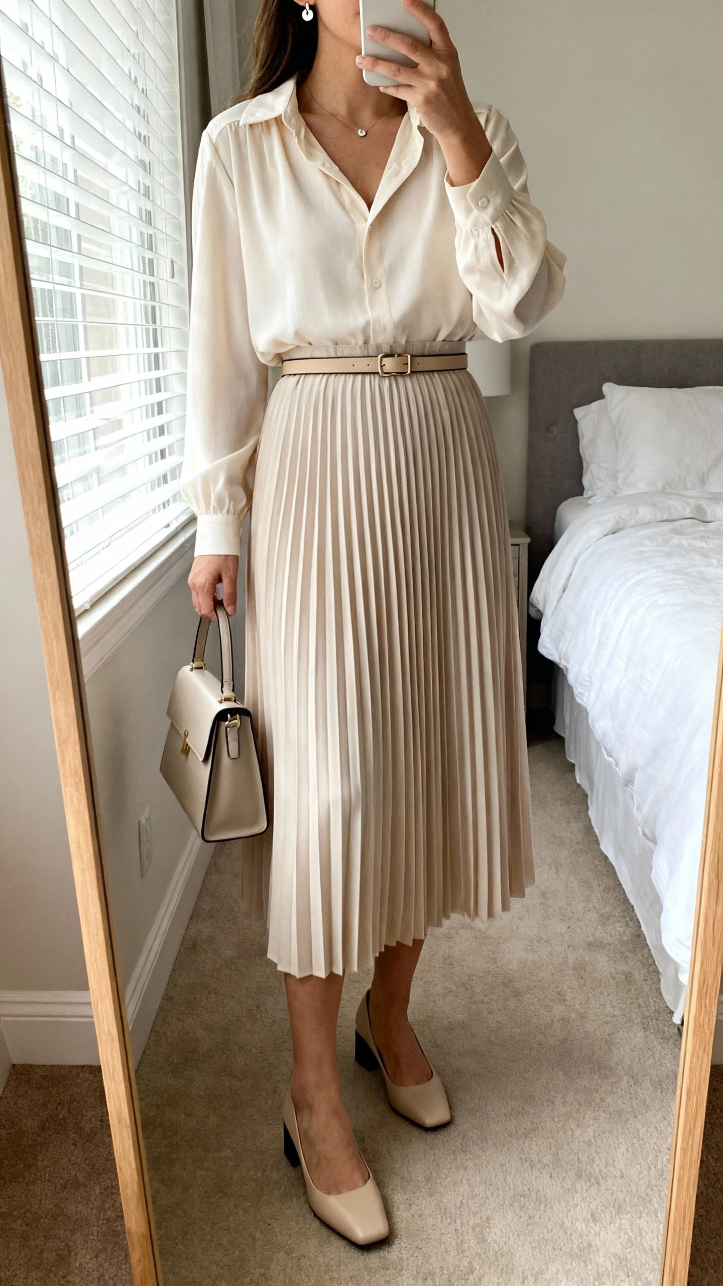 iPhone mirror selfie of a woman wearing a soft-pleated blouse tucked into a midi skirt with a slim belt, low block heels, delicate stud earrings, and a small structured handbag, face not visible, cropped at neck, bedroom mirror, natural window light, iPhone photo quality.