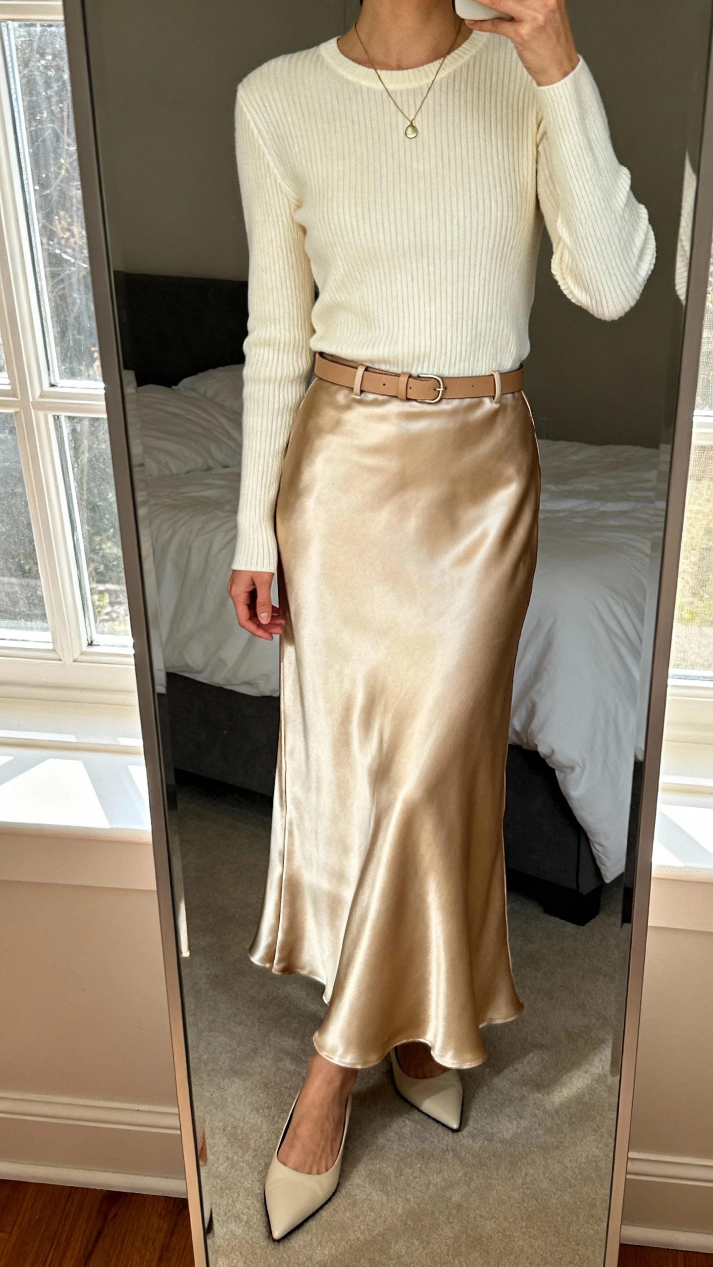 iPhone mirror selfie of a woman wearing a soft satin long skirt in champagne with a fitted ribbed knit top in cream, delicate pendant necklace, minimalist leather belt, and pointed-toe flats, face not visible, cropped at neck, bedroom mirror with natural window light, iPhone photo quality