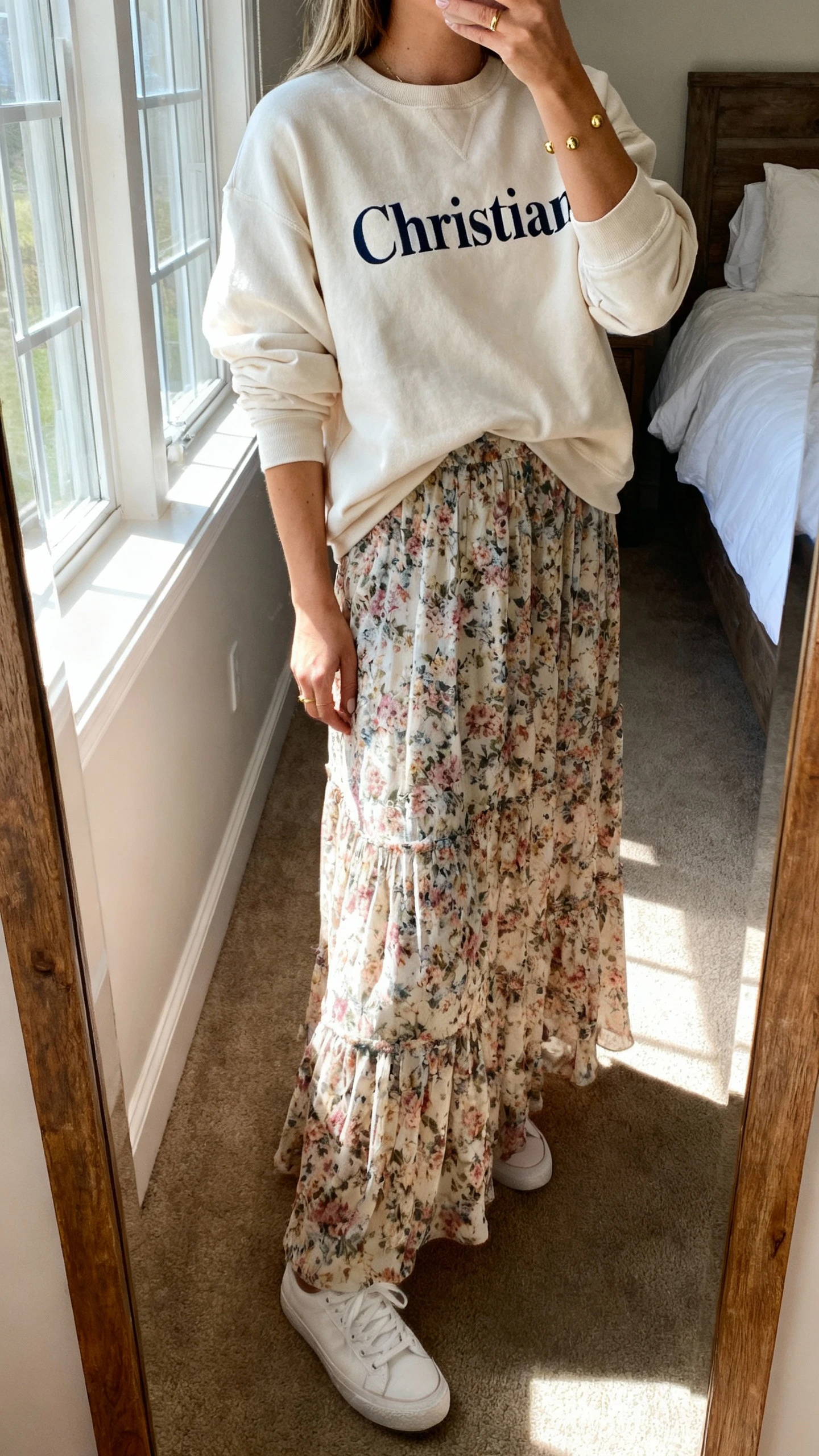 iPhone mirror selfie of a woman wearing a soft script Christian crewneck layered over a flowing floral maxi dress, with simple gold studs and white sneakers, face not visible, cropped at neck, bedroom mirror, morning window light, iPhone photo quality