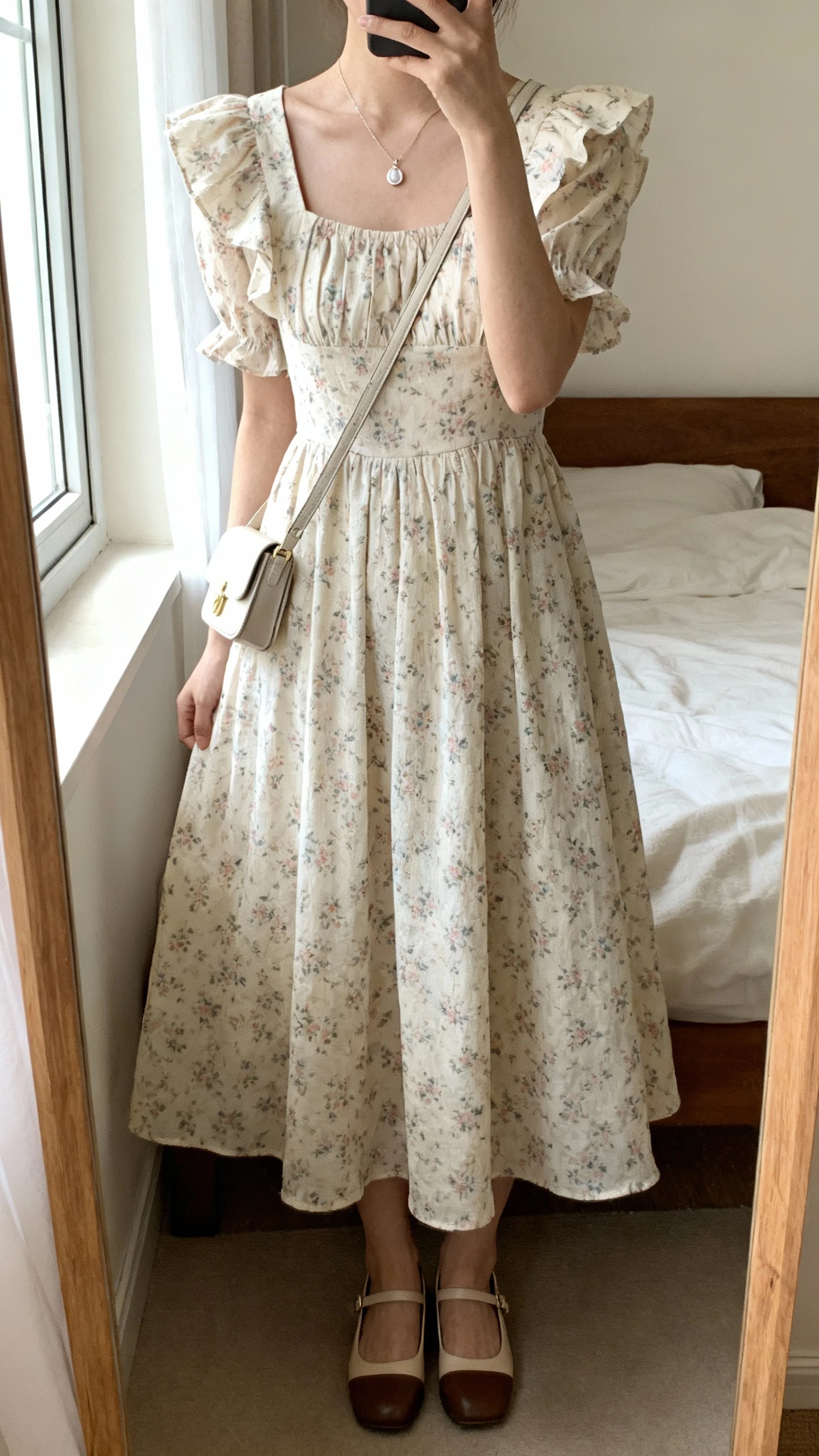 iPhone mirror selfie of a woman wearing a soft vintage-inspired floral tea dress with flutter sleeves, delicate pendant necklace, small crossbody bag, and low block-heel Mary Janes, face not visible, cropped at neck, bedroom mirror, morning window light, iPhone photo quality.