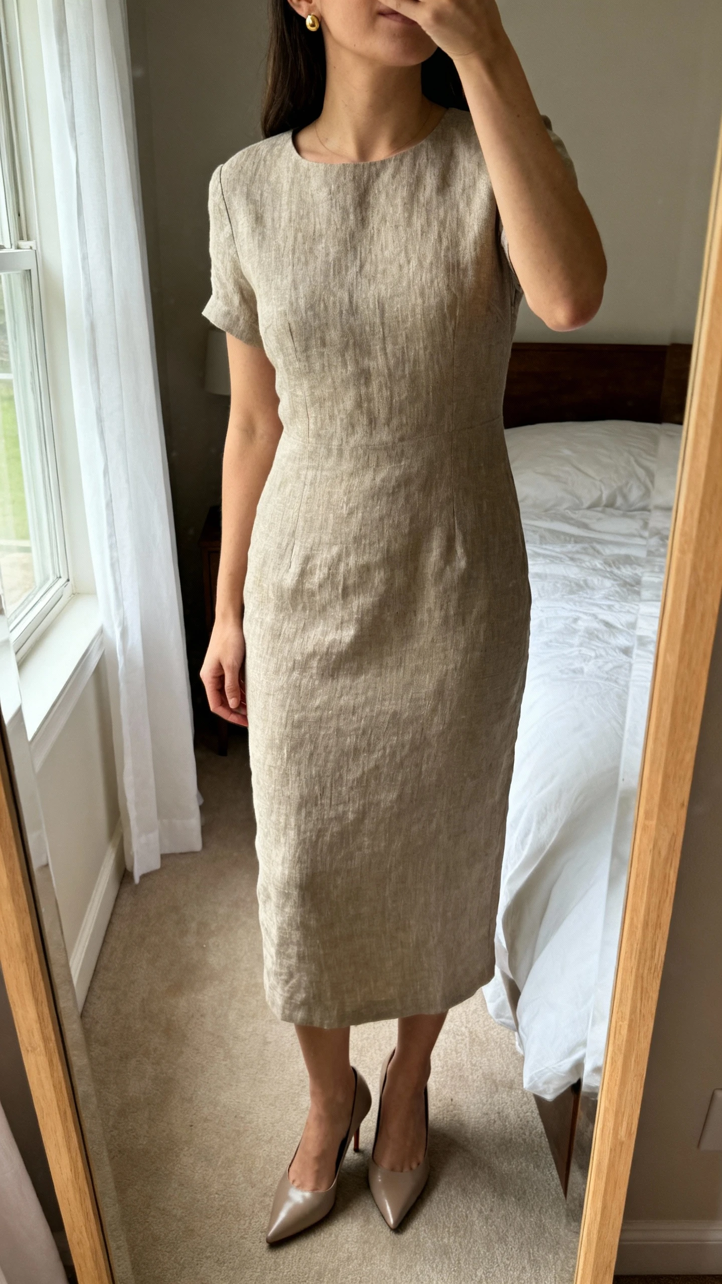 iPhone mirror selfie of a woman wearing a solid neutral linen sheath dress with a subtle quiet-luxury sheen, paired with minimal gold stud earrings and sleek kitten-heel pumps, face not visible, cropped at neck, bedroom mirror, soft morning window light, iPhone photo quality.