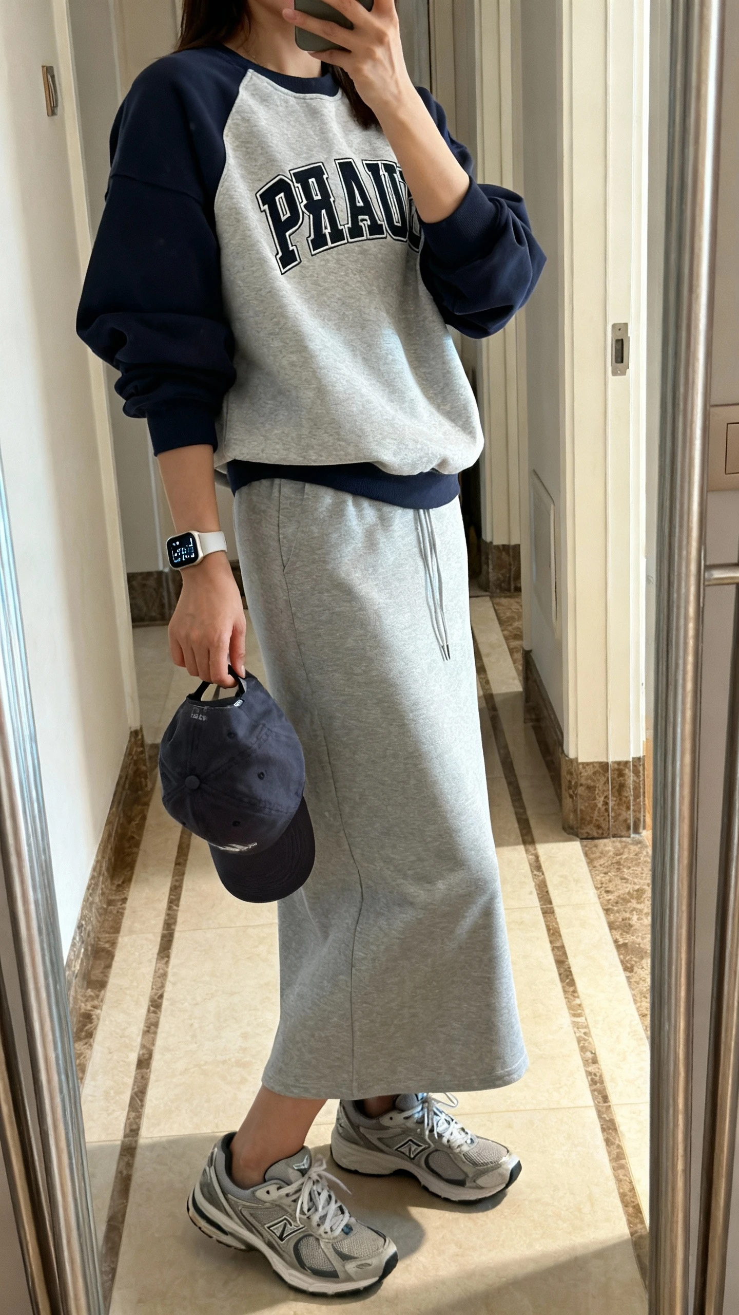 iPhone mirror selfie of a woman wearing a sporty praise crewneck with a jersey maxi skirt, baseball cap in hand, athletic sneakers, and a smartwatch, face not visible, side angle, entryway full-length mirror, indoor daylight, iPhone photo quality