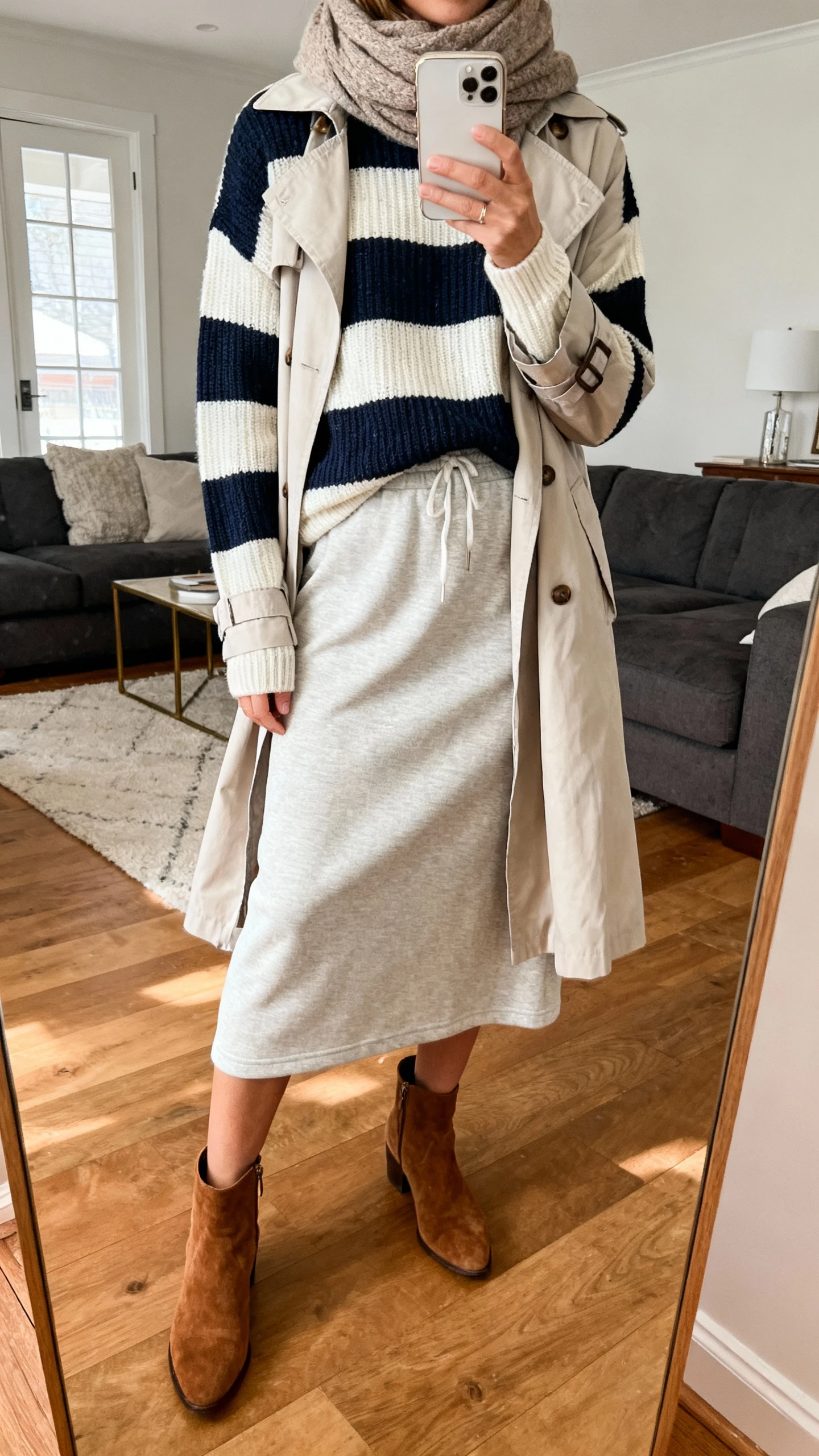 iPhone mirror selfie of a woman wearing a striped knit sweater over a soft jersey midi skirt, lightweight trench layered on top, suede ankle boots, cozy scarf, face not visible, cropped at neck, living room mirror, warm indoor lighting, iPhone photo quality.