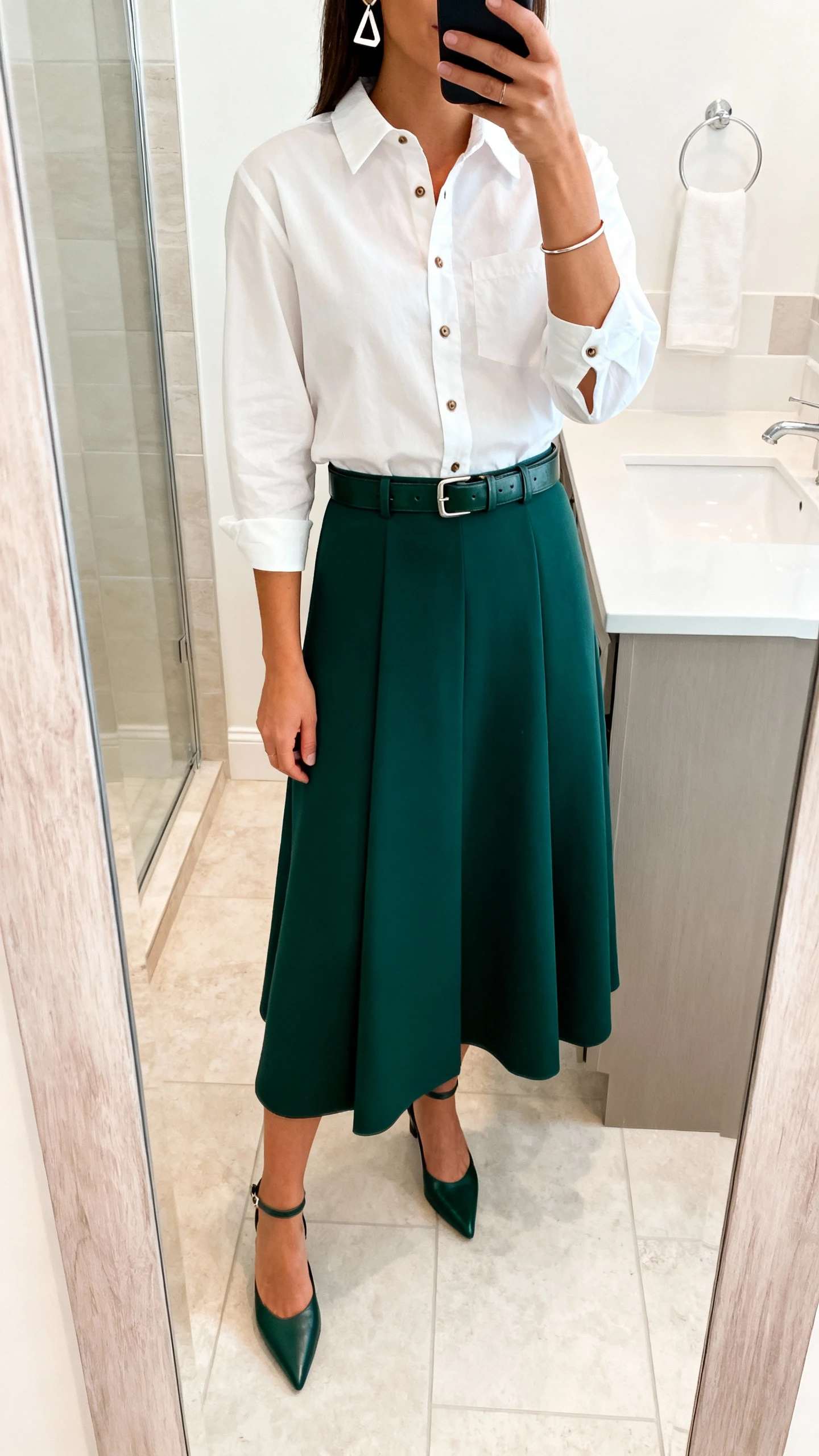 iPhone mirror selfie of a woman wearing a structured A-line maxi skirt in deep forest with a crisp white poplin button-up, sleek leather belt, pointed slingbacks, geometric earrings, and a slim cuff bracelet, face not visible, cropped at shoulders, bathroom mirror, bright indoor lighting, iPhone photo quality.