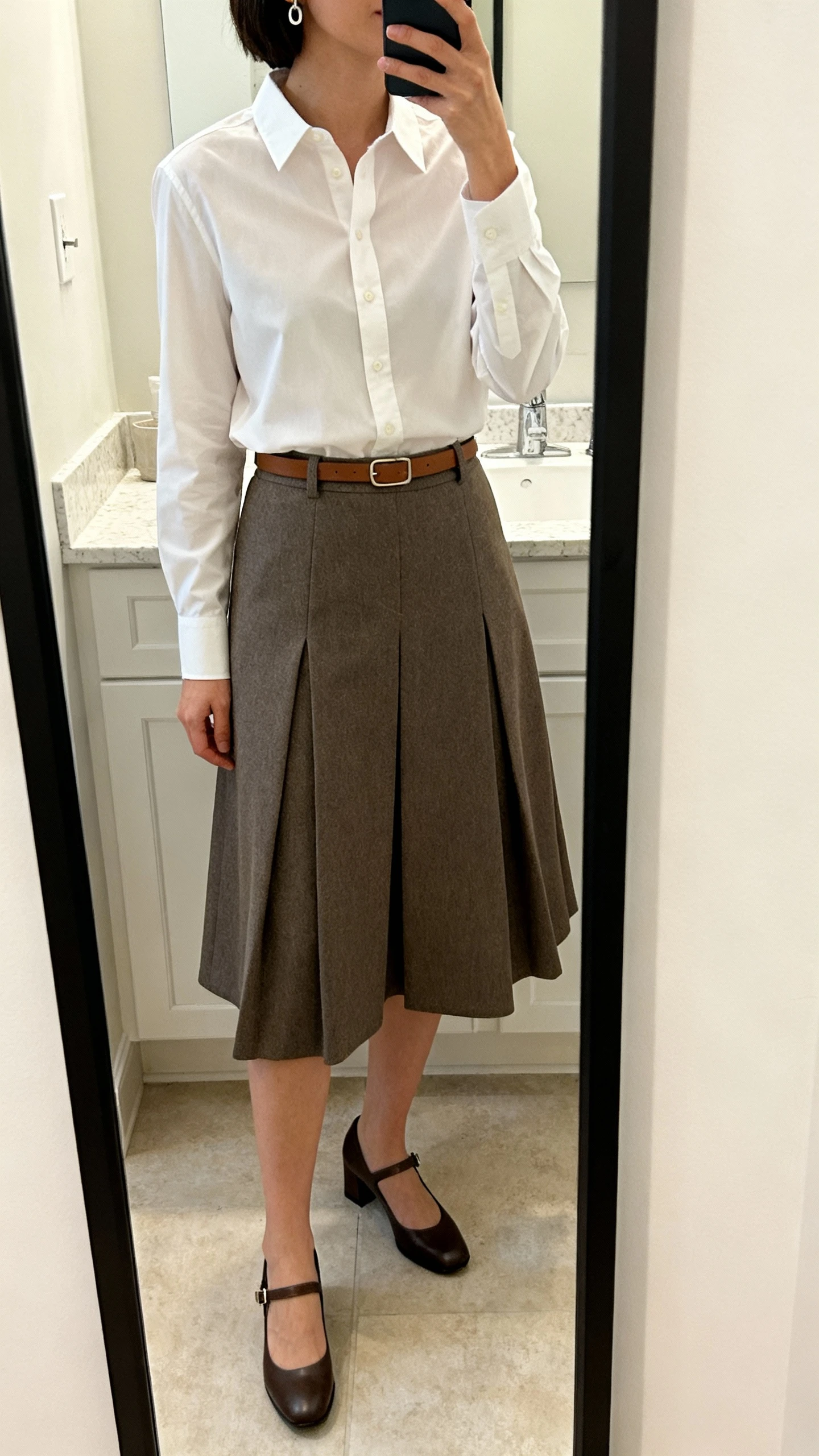 iPhone mirror selfie of a woman wearing a structured A-line midi skirt in twill, a crisp button-up shirt with a classic collar, low-heel Mary Janes, a minimal leather belt matching the shoes, and understated earrings; face not visible, cropped at shoulders, bathroom mirror, soft indoor lighting, iPhone photo quality.