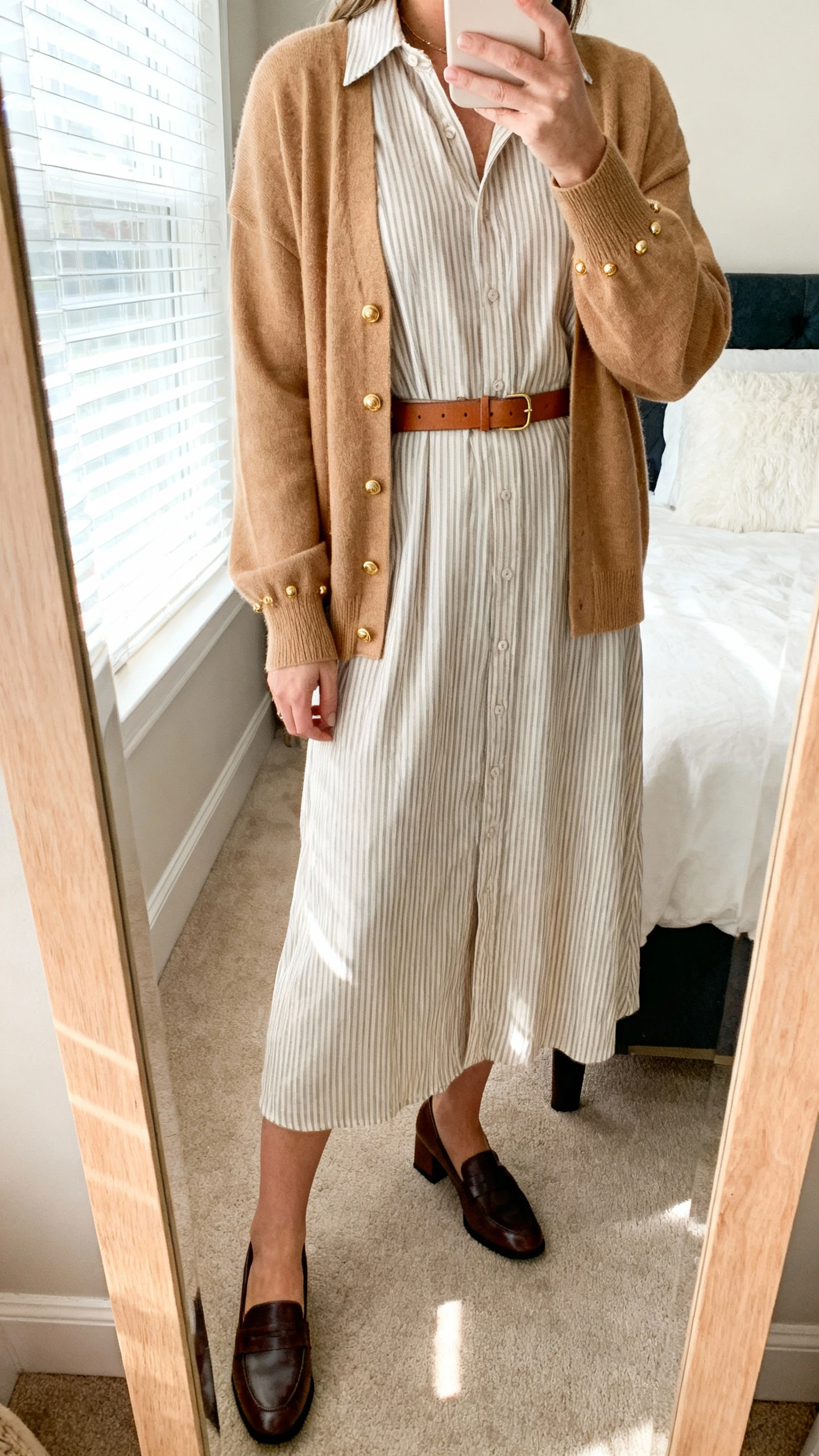 iPhone mirror selfie of a woman wearing a subtle striped midi dress with a belted camel cardigan, simple gold studs, slim leather belt, and low block-heel loafers, face not visible, cropped at neck, bedroom mirror, soft morning window light, iPhone photo quality.