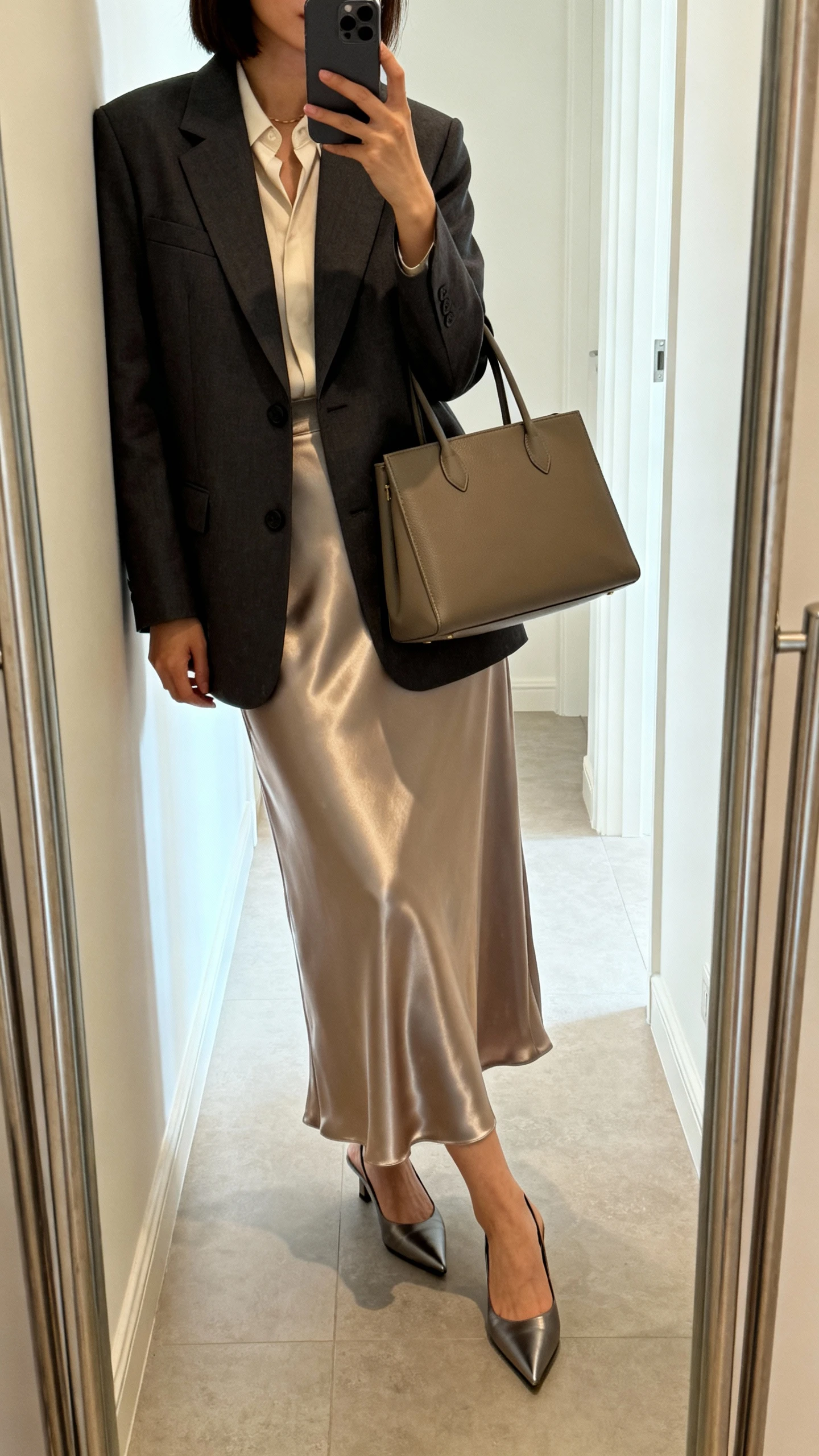 iPhone mirror selfie of a woman wearing a tailored blazer over a satin midi-maxi skirt, minimalist blouse, structured tote, and pointed-toe heels, face not visible, side angle at full-length mirror in a hallway, bright indoor lighting, iPhone photo quality.