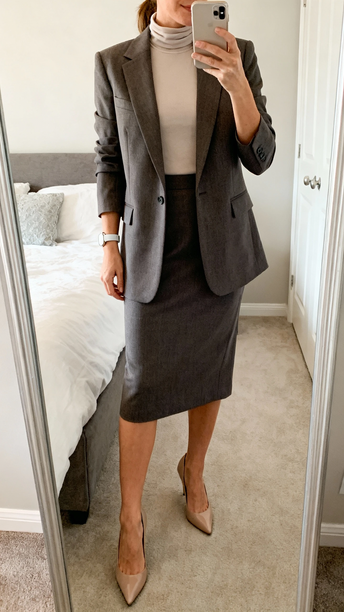iPhone mirror selfie of a woman wearing a tailored suiting skirt set (knee-length skirt and matching blazer) over a high-neck shell top, classic pumps and a simple watch, face not visible, cropped at shoulders, bedroom mirror, neutral indoor lighting, iPhone photo quality.