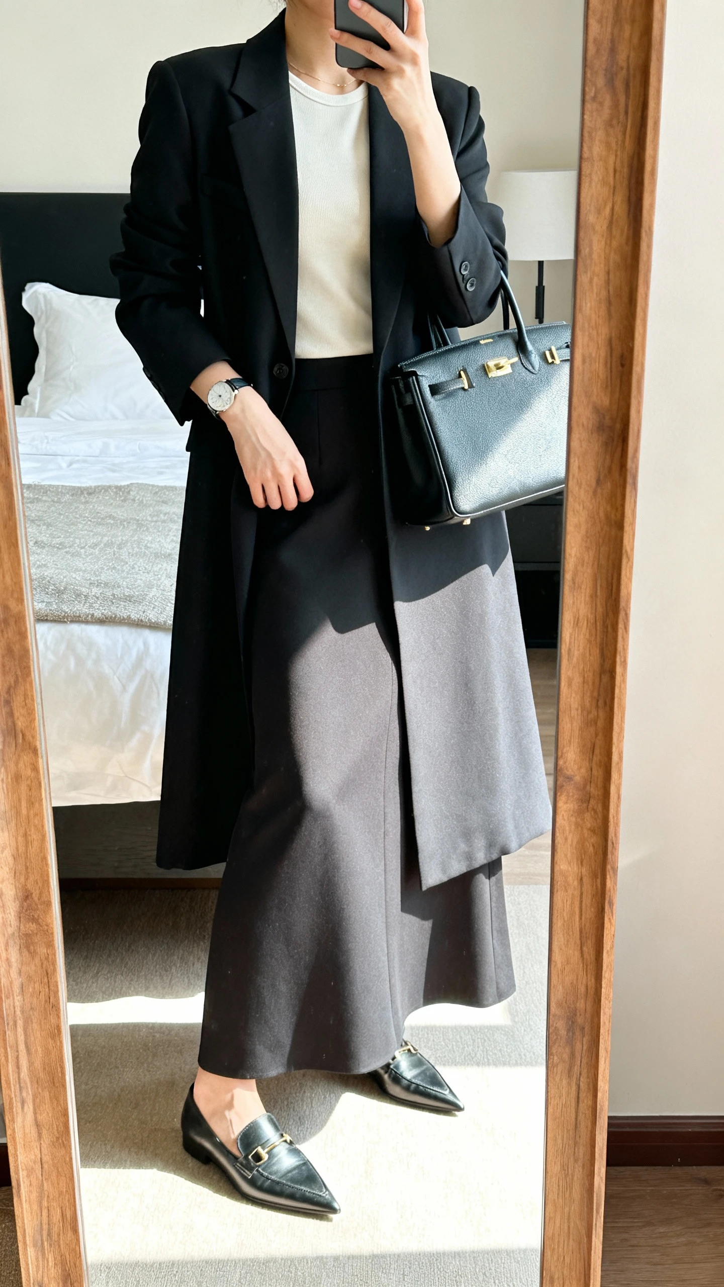 iPhone mirror selfie of a woman wearing a tailored maxi skirt with a longline blazer over a modest shell top, pointed-toe loafers, minimalist watch, and a structured handbag, face not visible, cropped at shoulders, bedroom mirror, bright indoor lighting, iPhone photo quality.