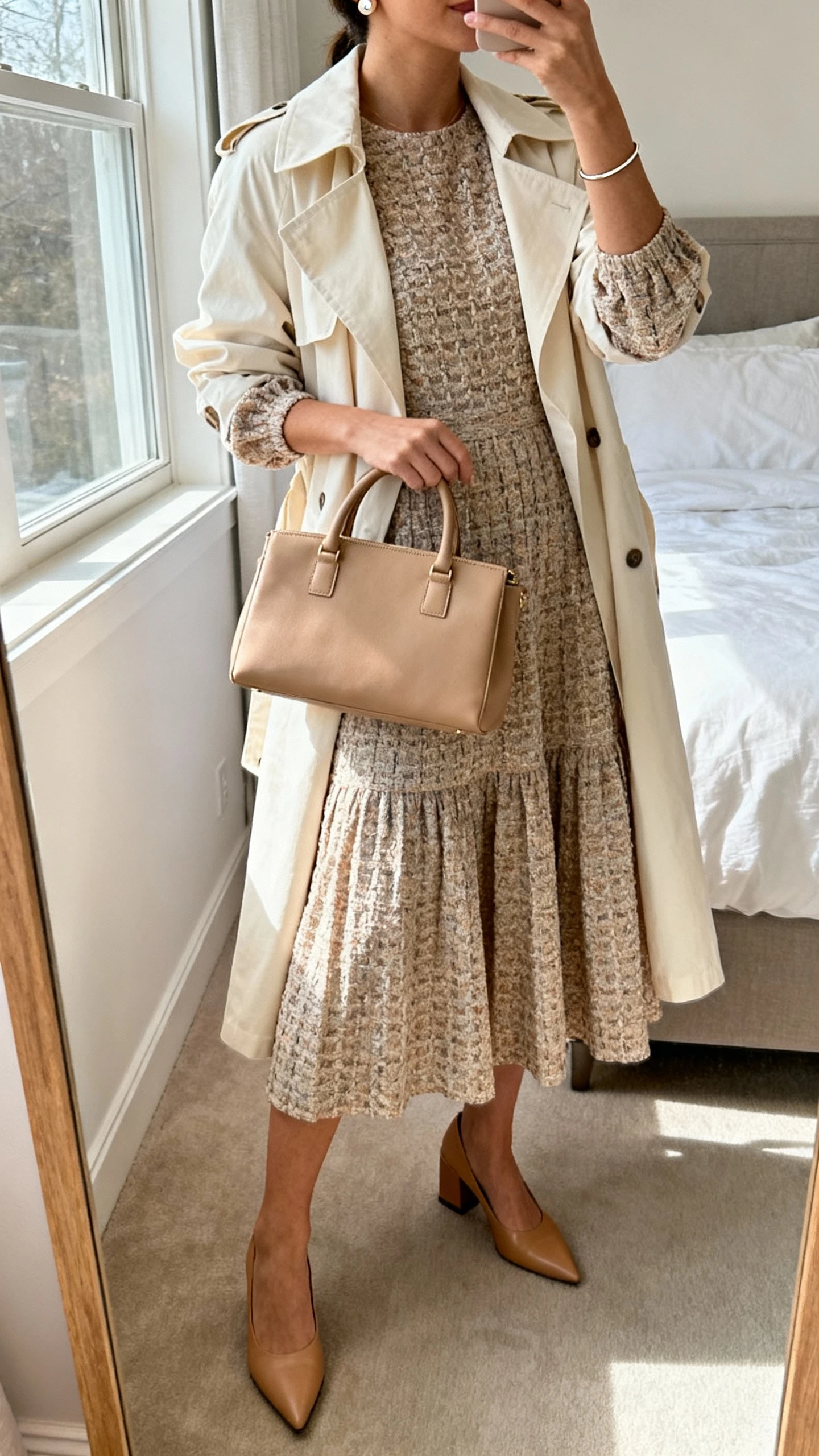 iPhone mirror selfie of a woman wearing a textured jacquard midi dress with elbow-length sleeves and a flowy A-line skirt, a lightweight cream trench, pointed-toe block heels in tan, a structured top-handle bag in a coordinating neutral, delicate stud earrings, and a slim bangle; face not visible, cropped at neck, bedroom mirror, natural window light, iPhone photo quality.