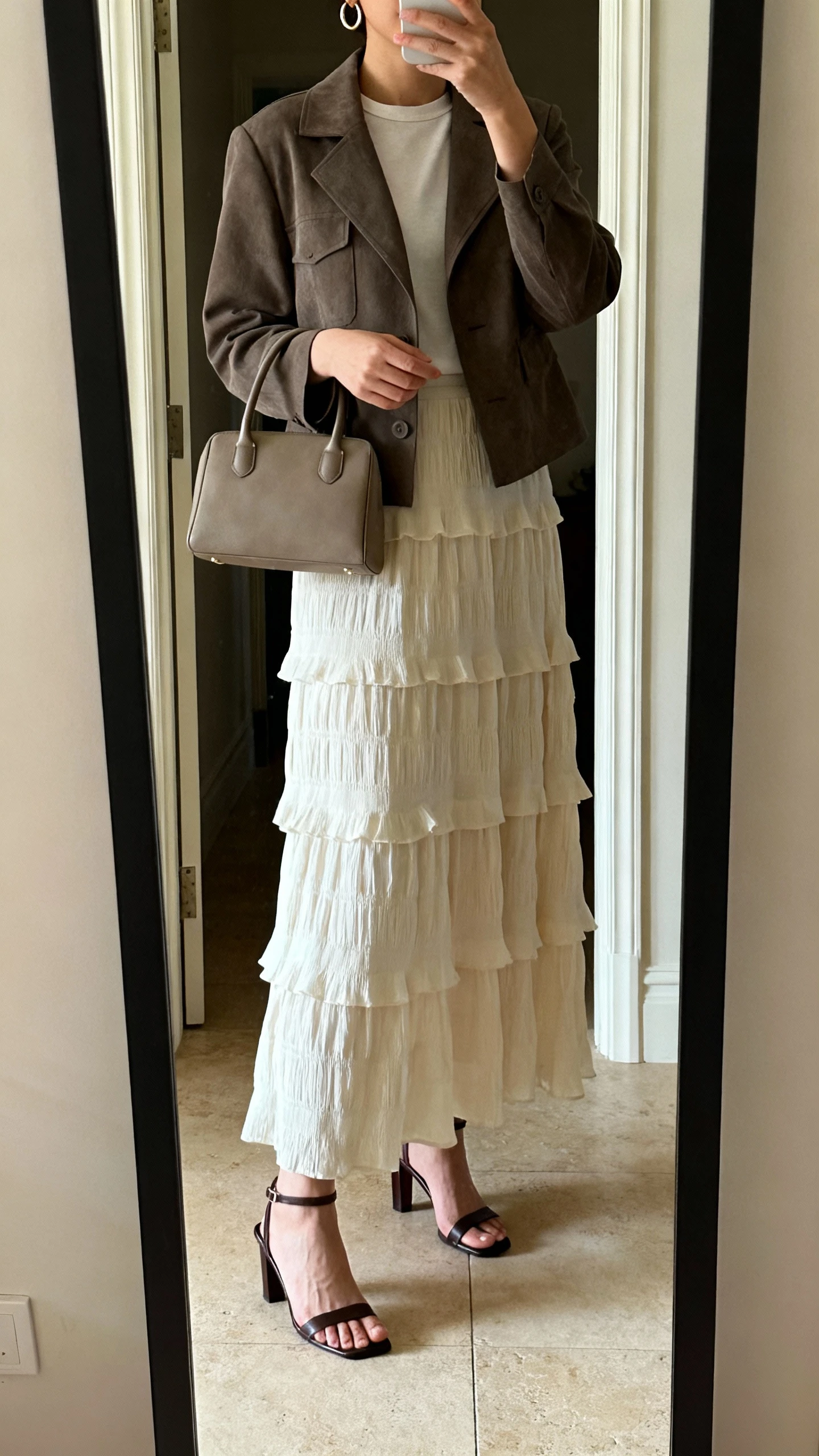 iPhone mirror selfie of a woman wearing a tiered maxi skirt with a structured jacket over a simple top, heeled sandals, a top-handle bag, and subtle hoop earrings, face not visible, cropped at neck, entryway full-length mirror, warm indoor lighting, iPhone photo quality.
