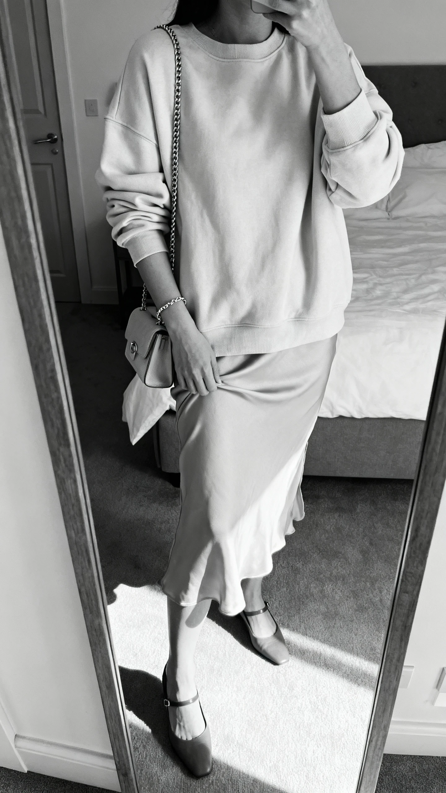 iPhone mirror selfie of a woman wearing a tonal crewneck layered over a matching slip dress, monochrome palette, delicate chain bracelet, small shoulder bag, and low slingback heels; face not visible, cropped at shoulders, bedroom mirror, gentle morning light, iPhone photo quality.